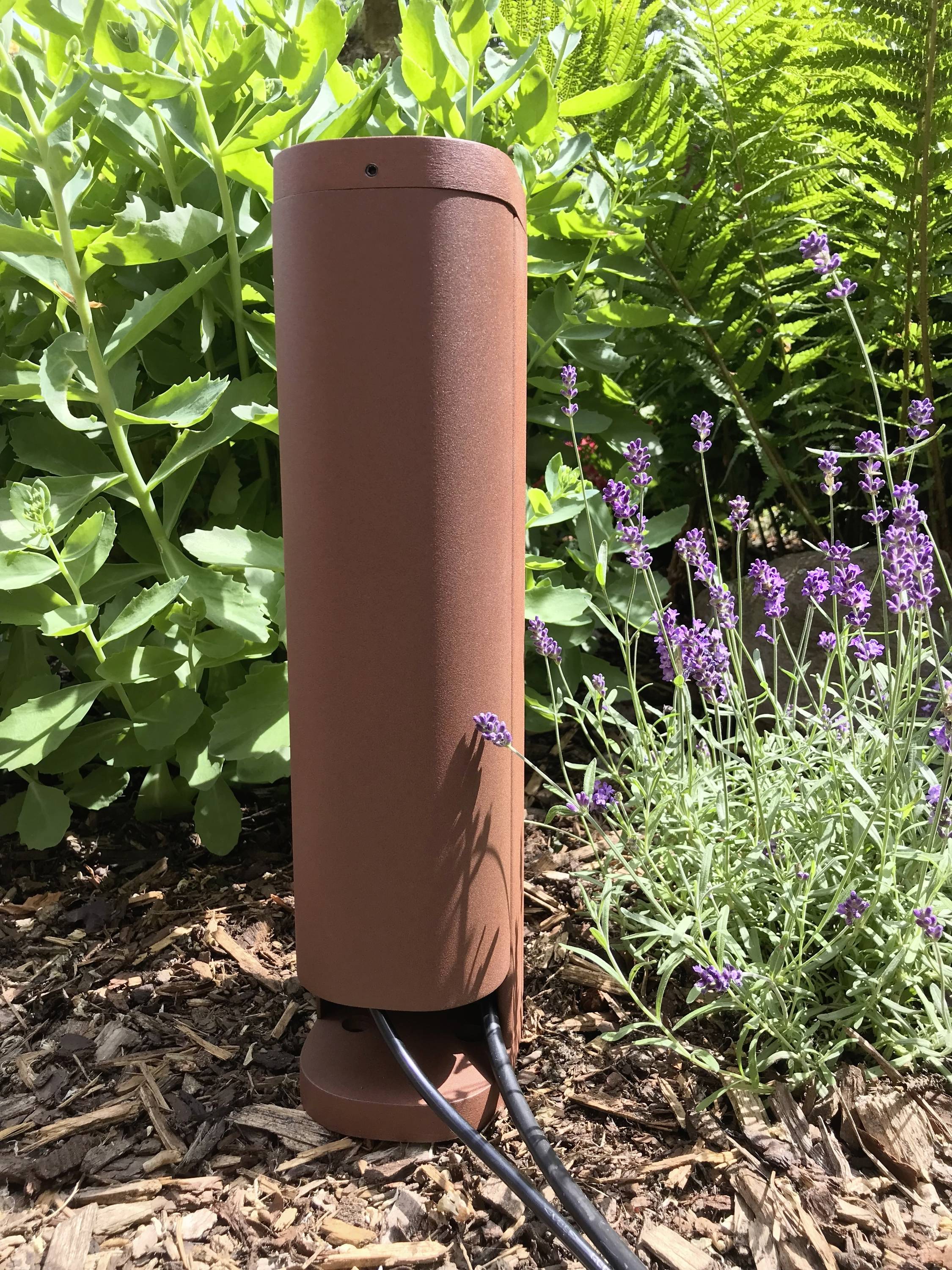 Une lampe de jardin cylindrique marron est placée dans un massif de lavande et de plantes vertes. Des câbles partent de la lampe.