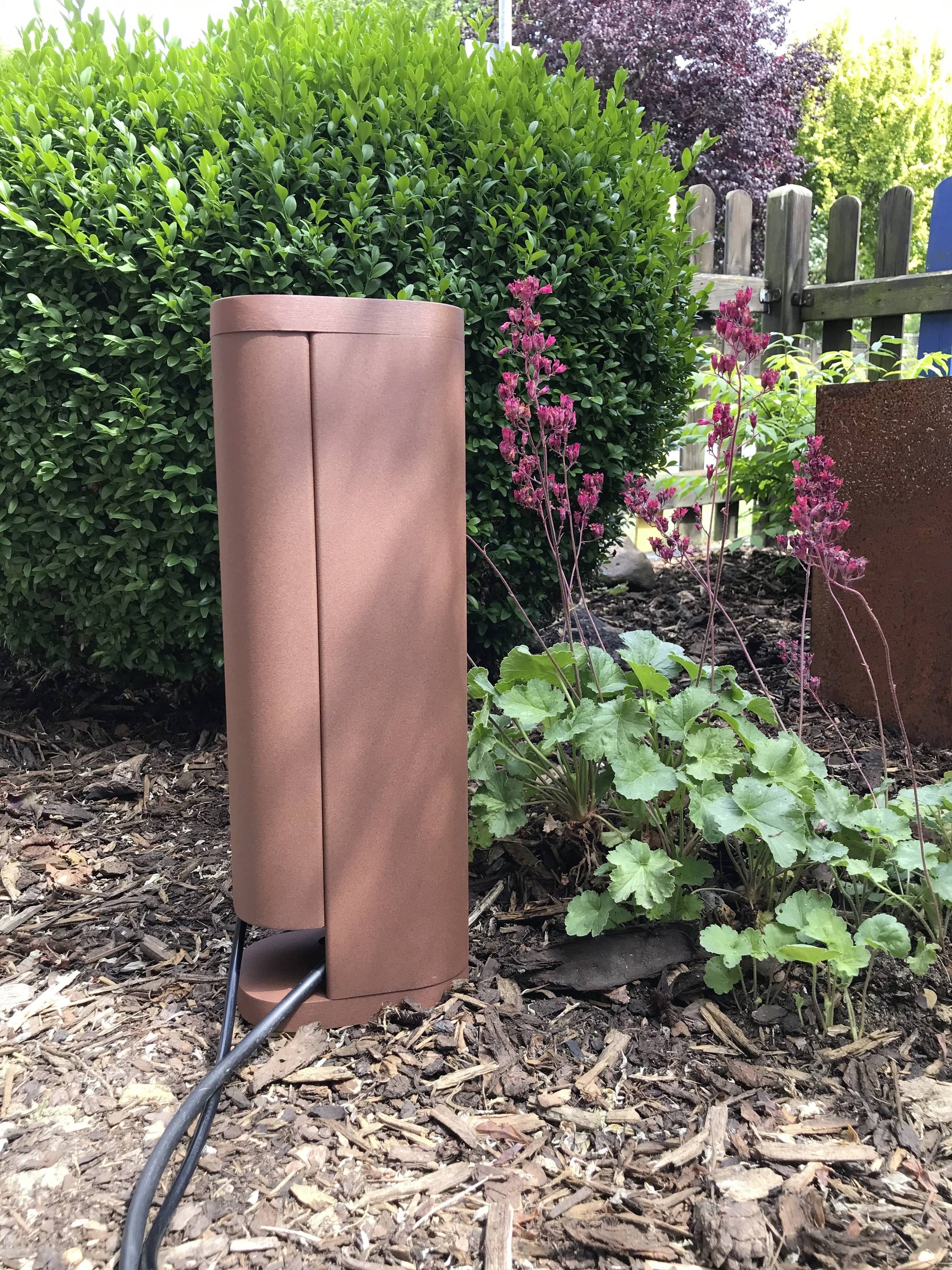 Une fontaine en bronze dans un jardin, entourée de plantes vertes et rouges, posée sur un lit de paillis, raccordée à un câble.