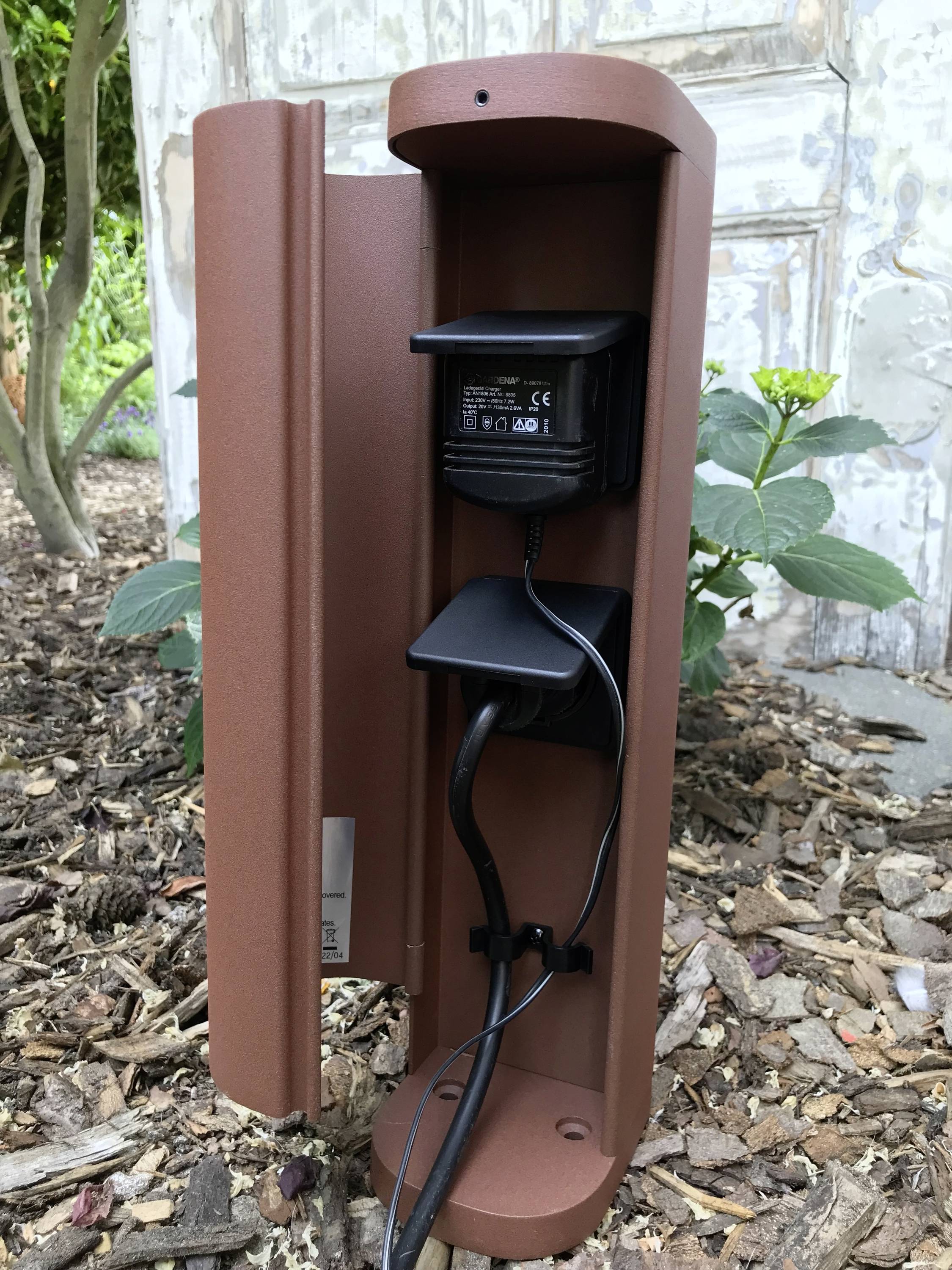 Une colonne de prise électrique marron pour extérieur, avec une fiche câblée, est plantée dans la terre devant un mur en bois, entourée de plantes.