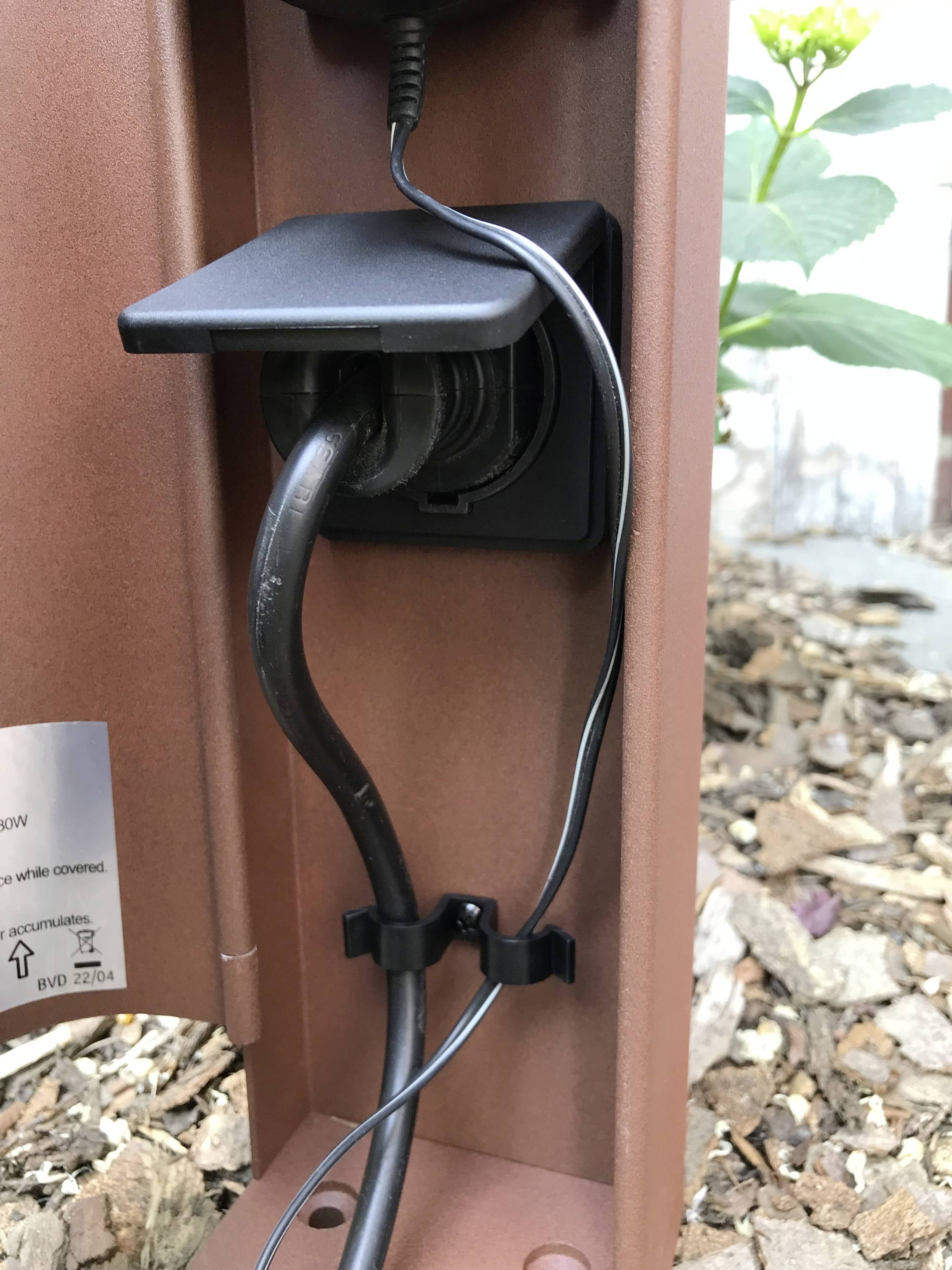 Une prise électrique extérieure avec un couvercle ouvert montre deux câbles branchés. En arrière-plan, on peut voir des plantes et des copeaux de bois.