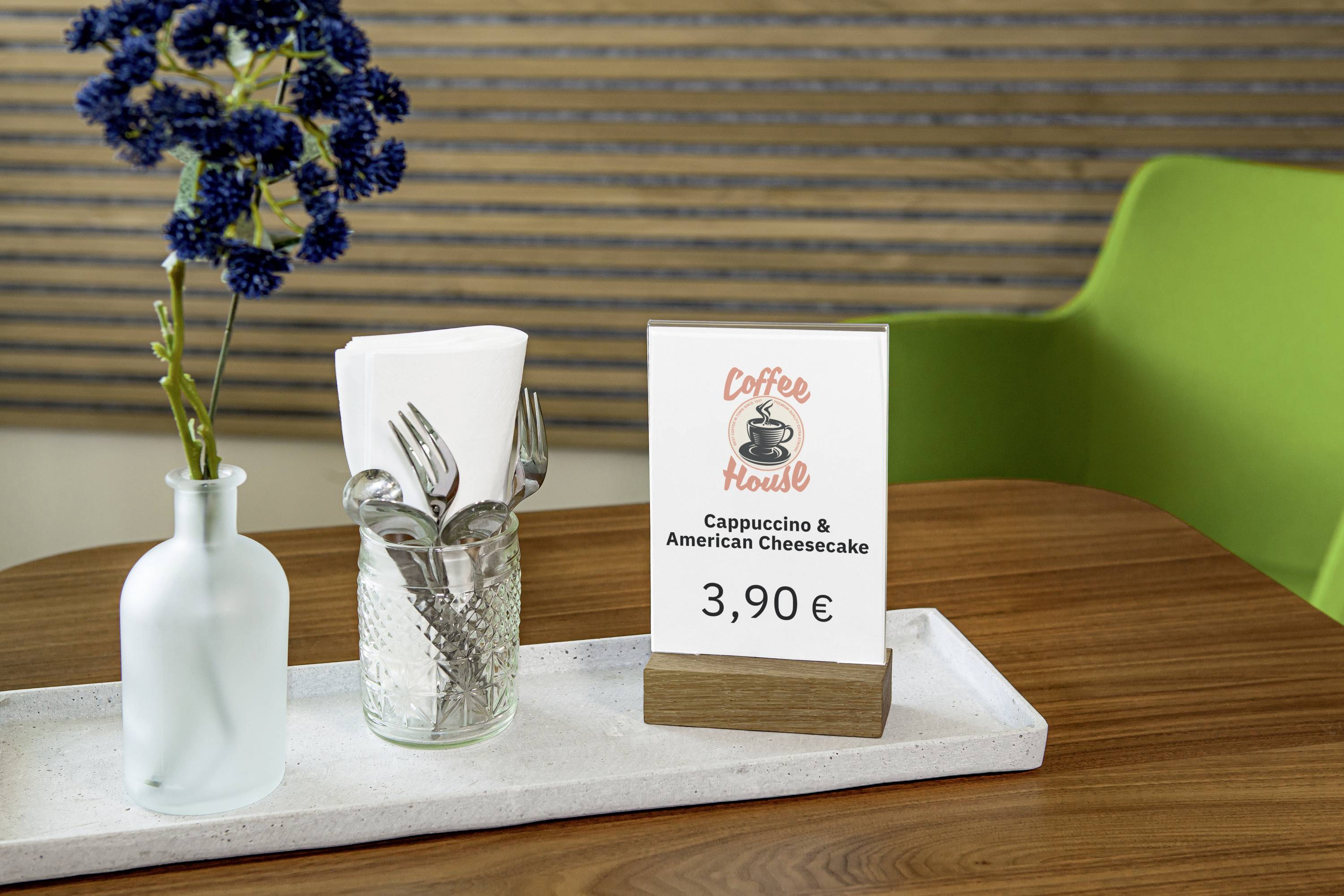 Une table avec un verre et des couverts, un vase avec des fleurs et un support annonçant un 'Cappuccino & American Cheesecake' pour 3,90 €.