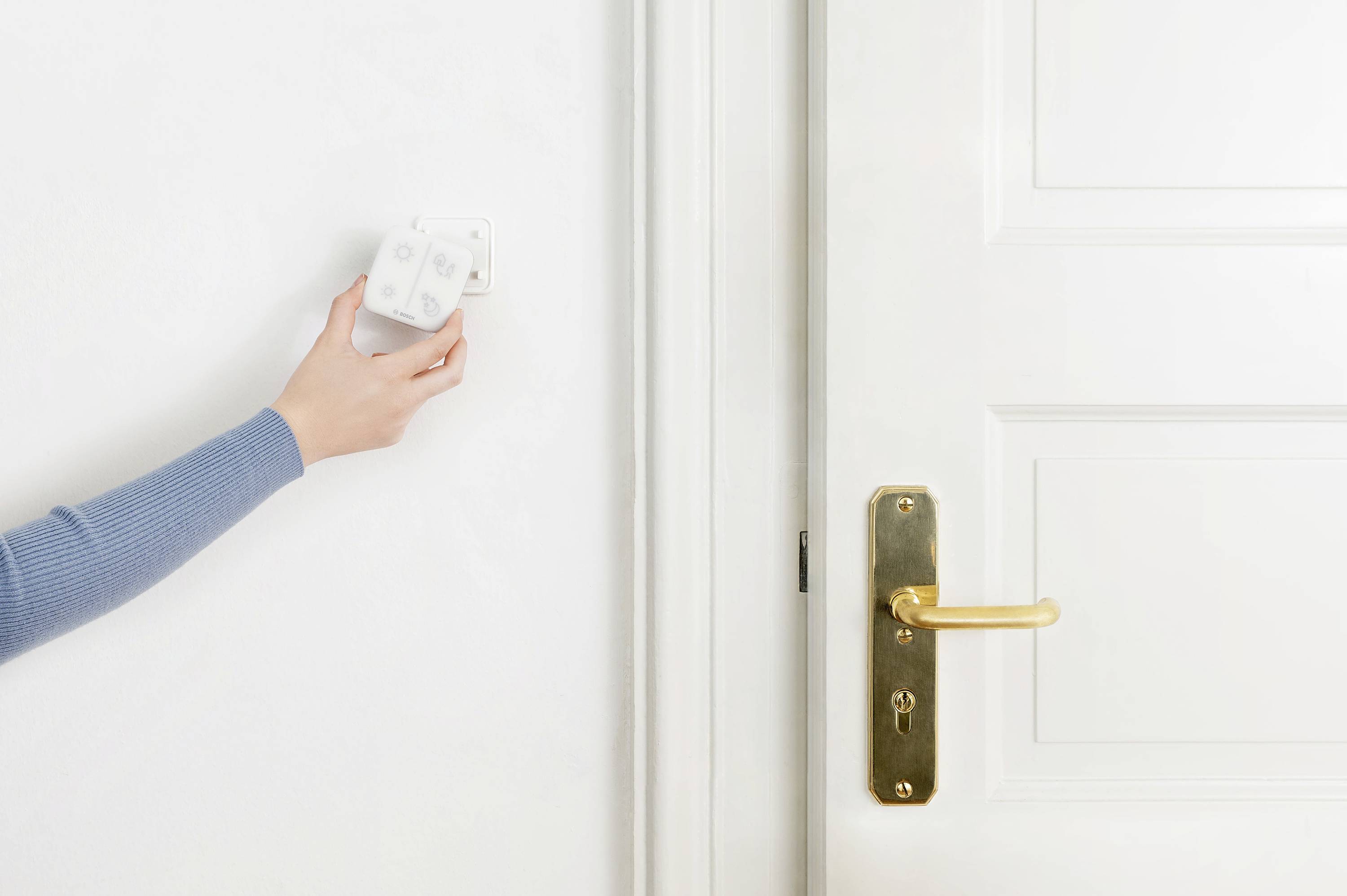 Une main ajuste un thermostat sur le mur à côté d'une porte blanche avec une poignée dorée.
