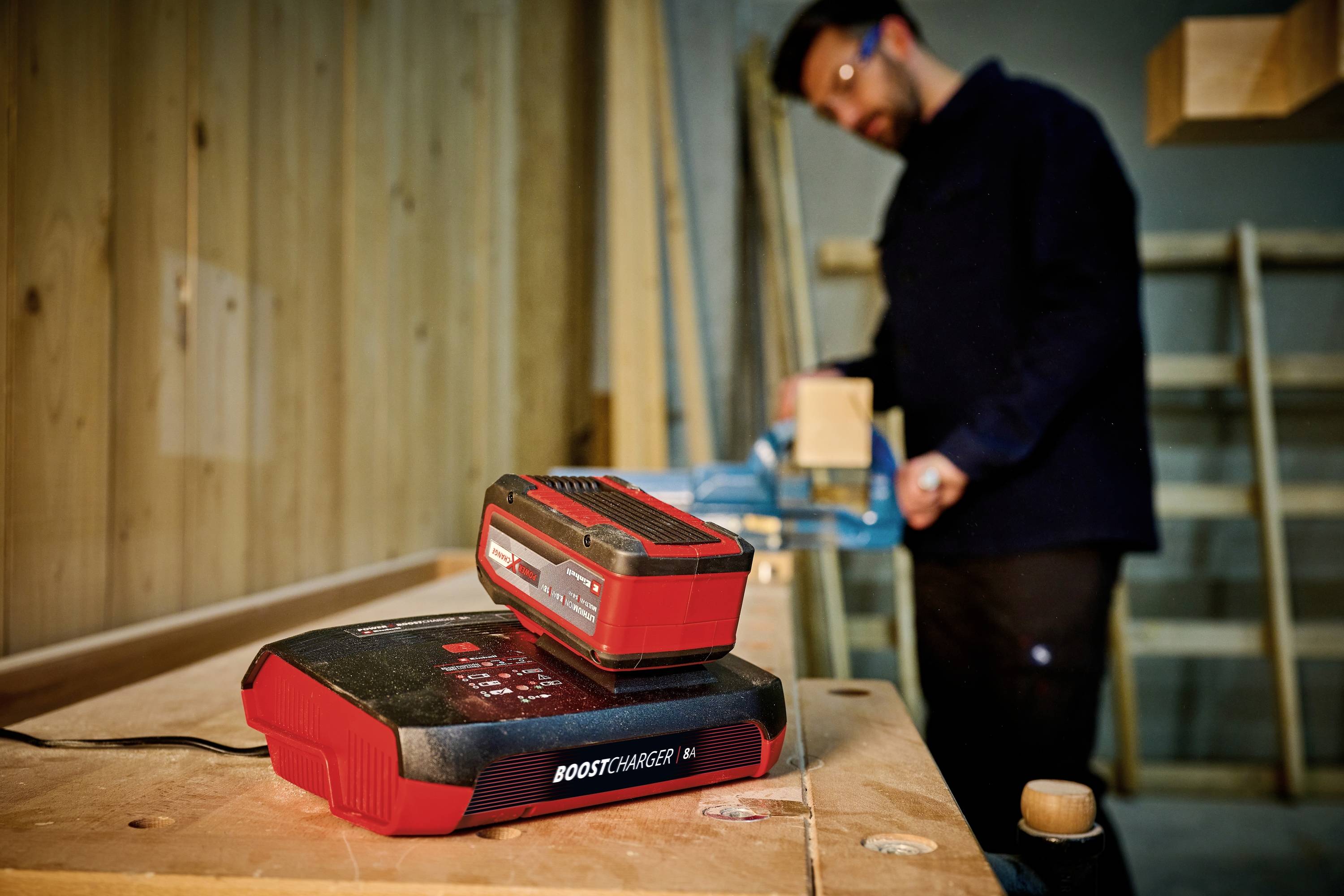 Un chargeur rouge avec une batterie installée est placé au premier plan sur une table, tandis qu'une personne travaille le bois en arrière-plan.