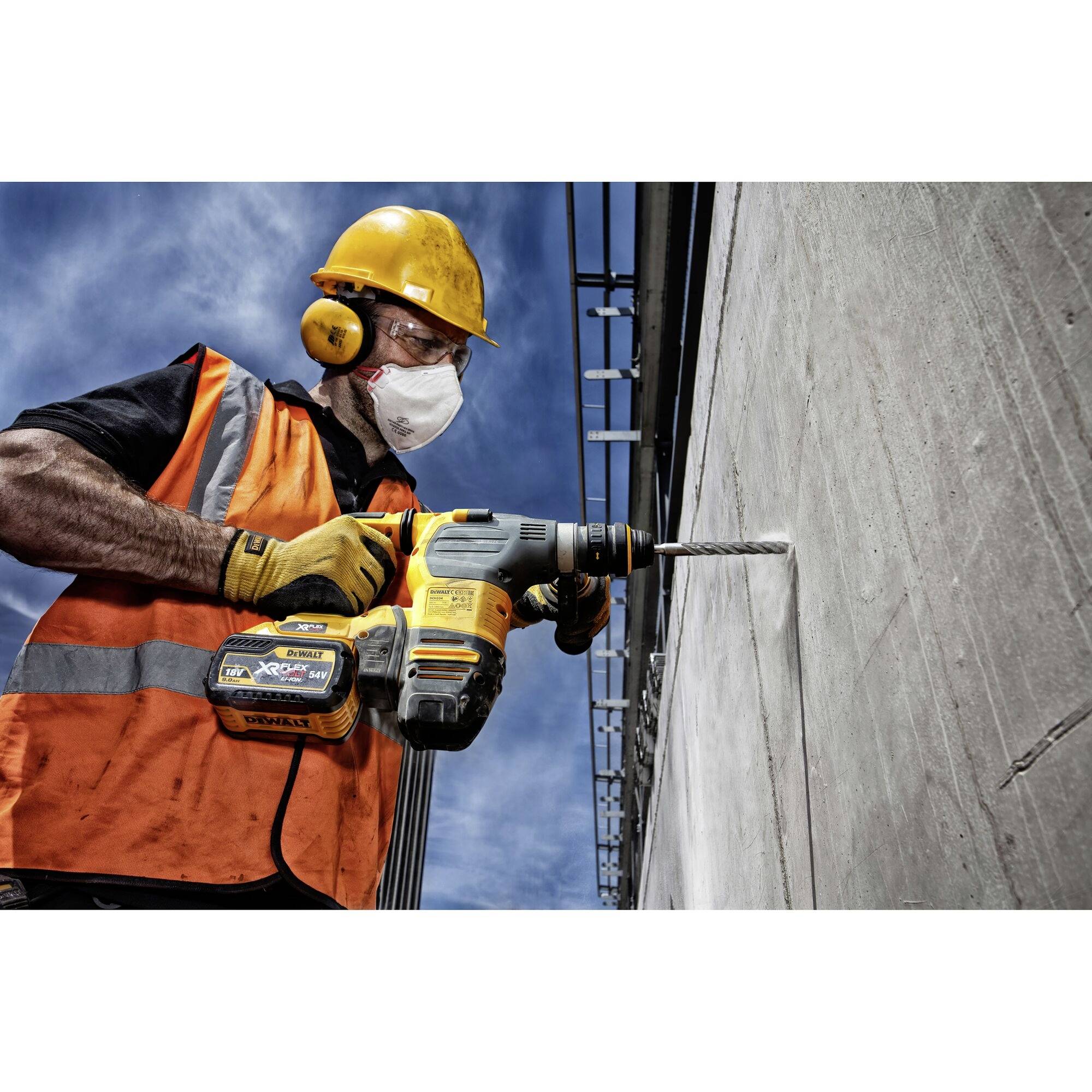 Un ouvrier portant un gilet orange et un casque de protection fore un trou dans un mur en béton à l'aide d'une perceuse électrique.