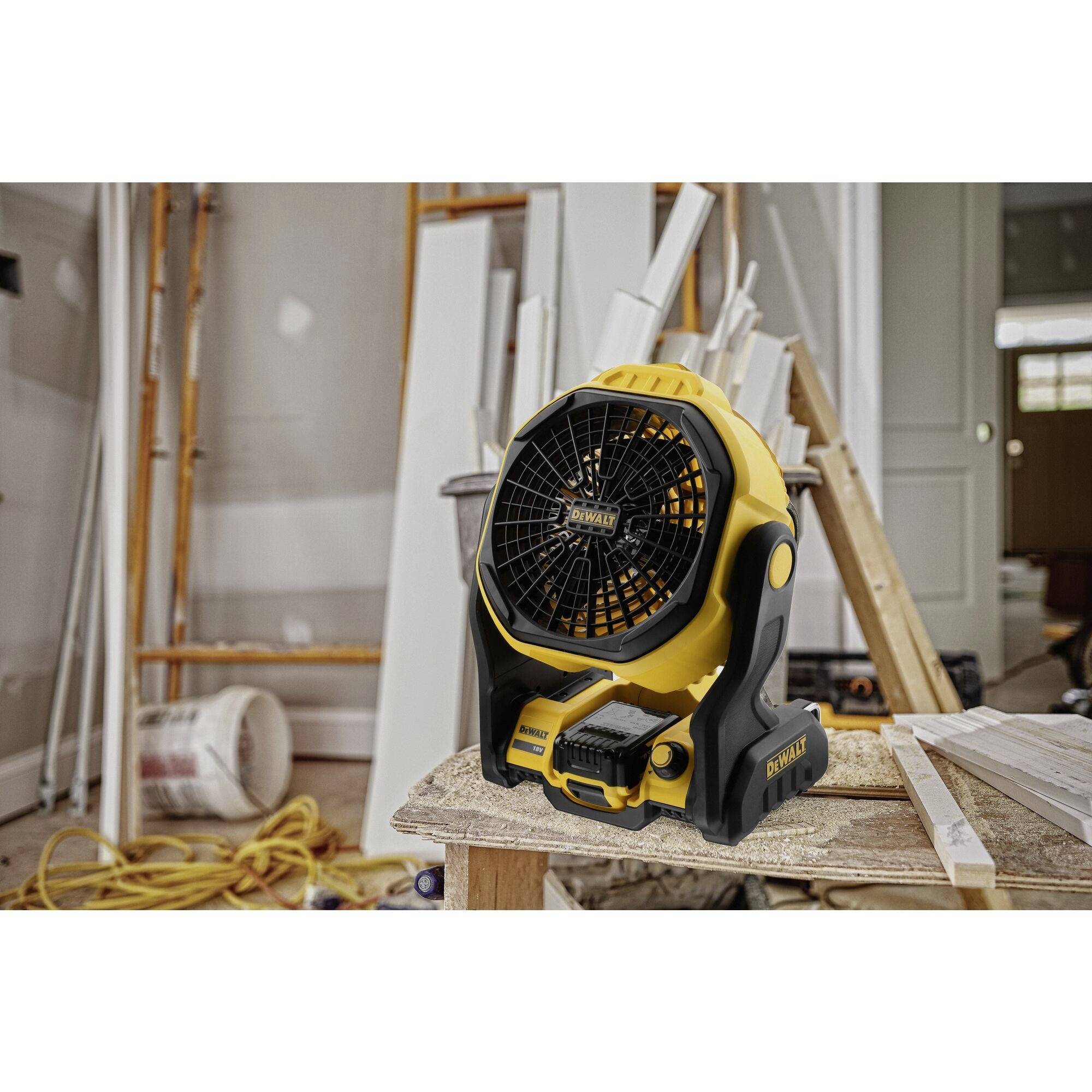 Un ventilateur jaune est posé sur une table dans un atelier. À l'arrière-plan, on peut voir des matériaux de construction et des échafaudages.