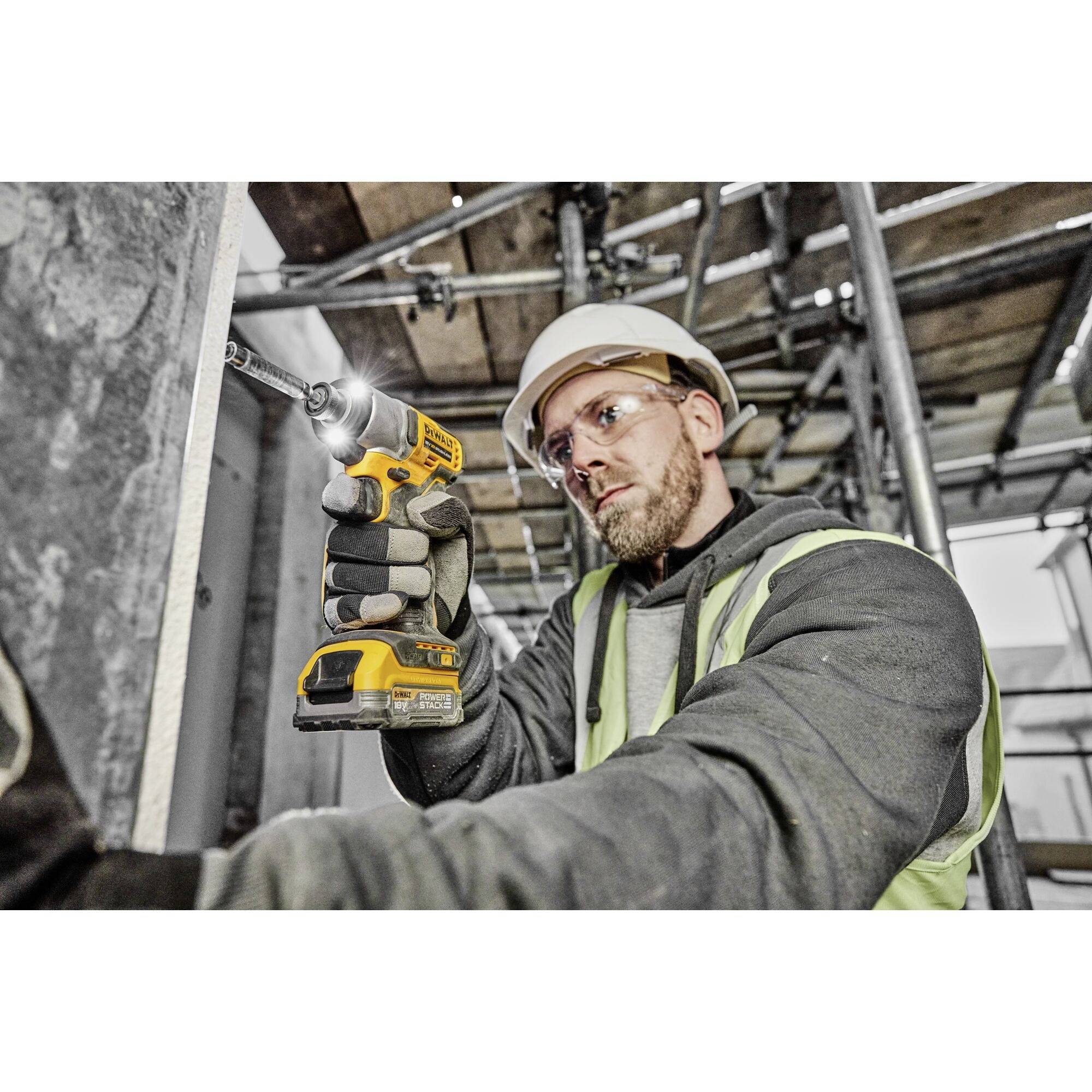 Un ouvrier portant un casque de chantier et un gilet de sécurité fore du béton avec une perceuse sans fil sur un chantier.