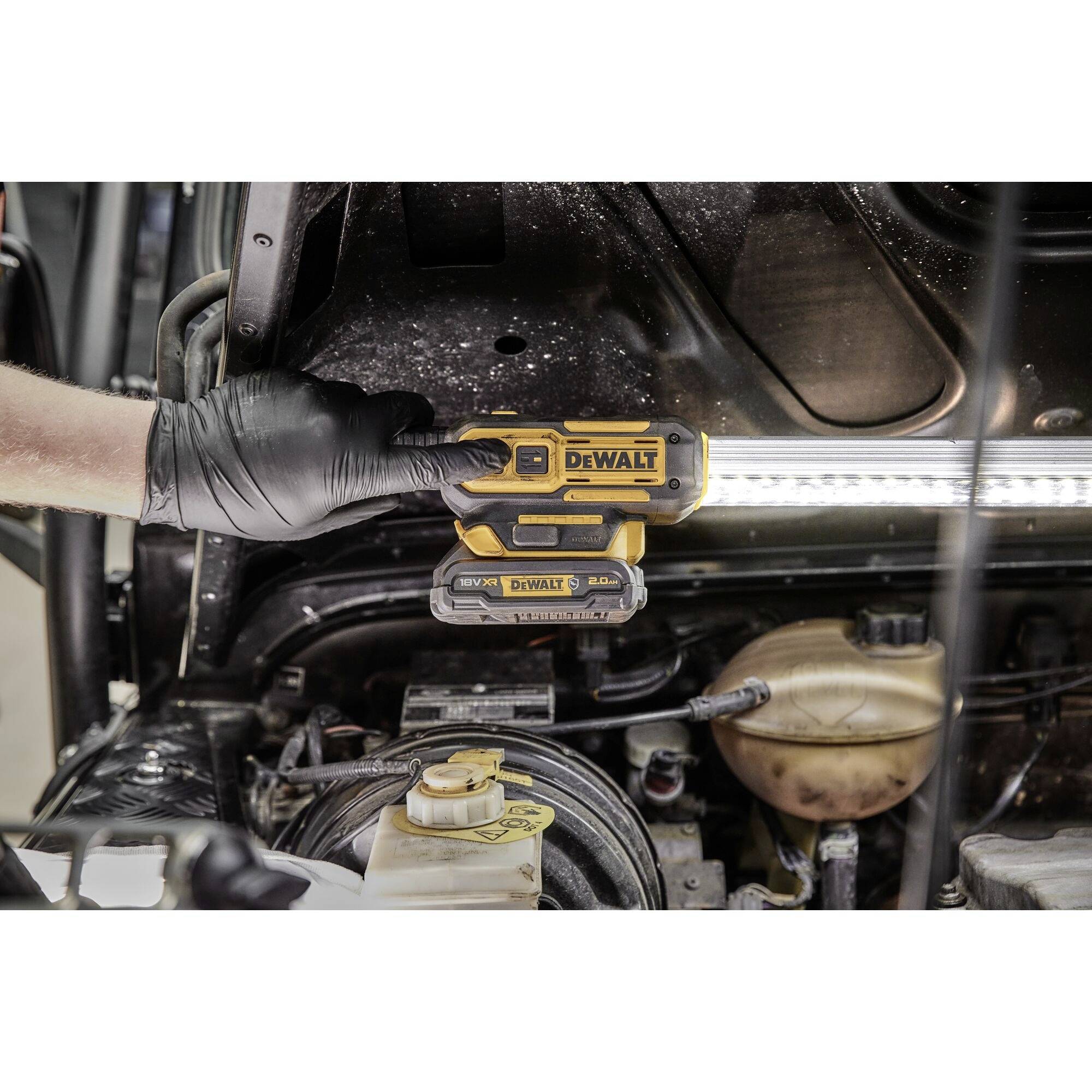 Une personne portant des gants noirs tient un outil DeWalt sous le capot d'un véhicule afin d'éclairer le compartiment moteur.