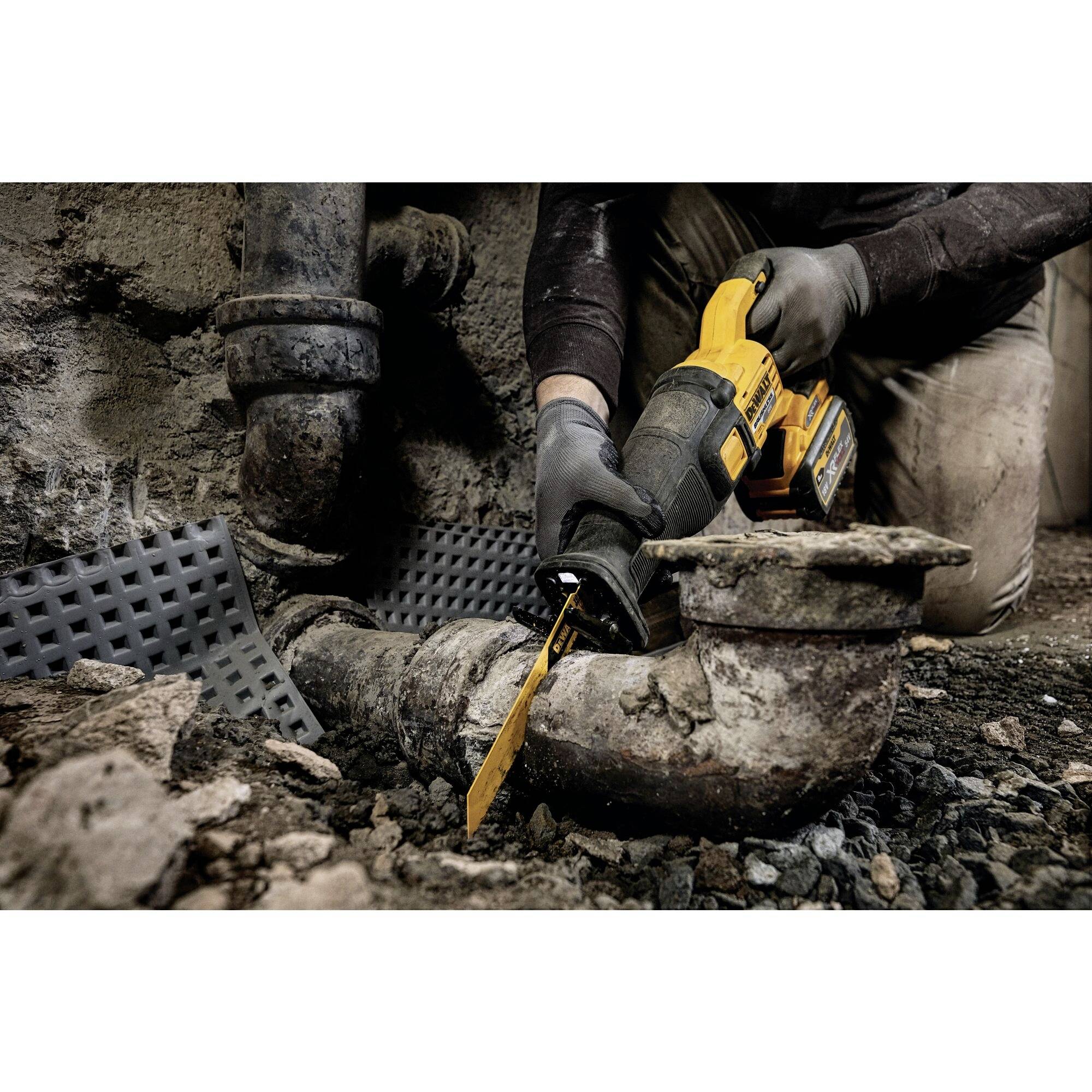Une personne coupe des tuyaux anciens et rouillés avec une scie électrique dans une cave. Travaille de manière concentrée, portant des gants.