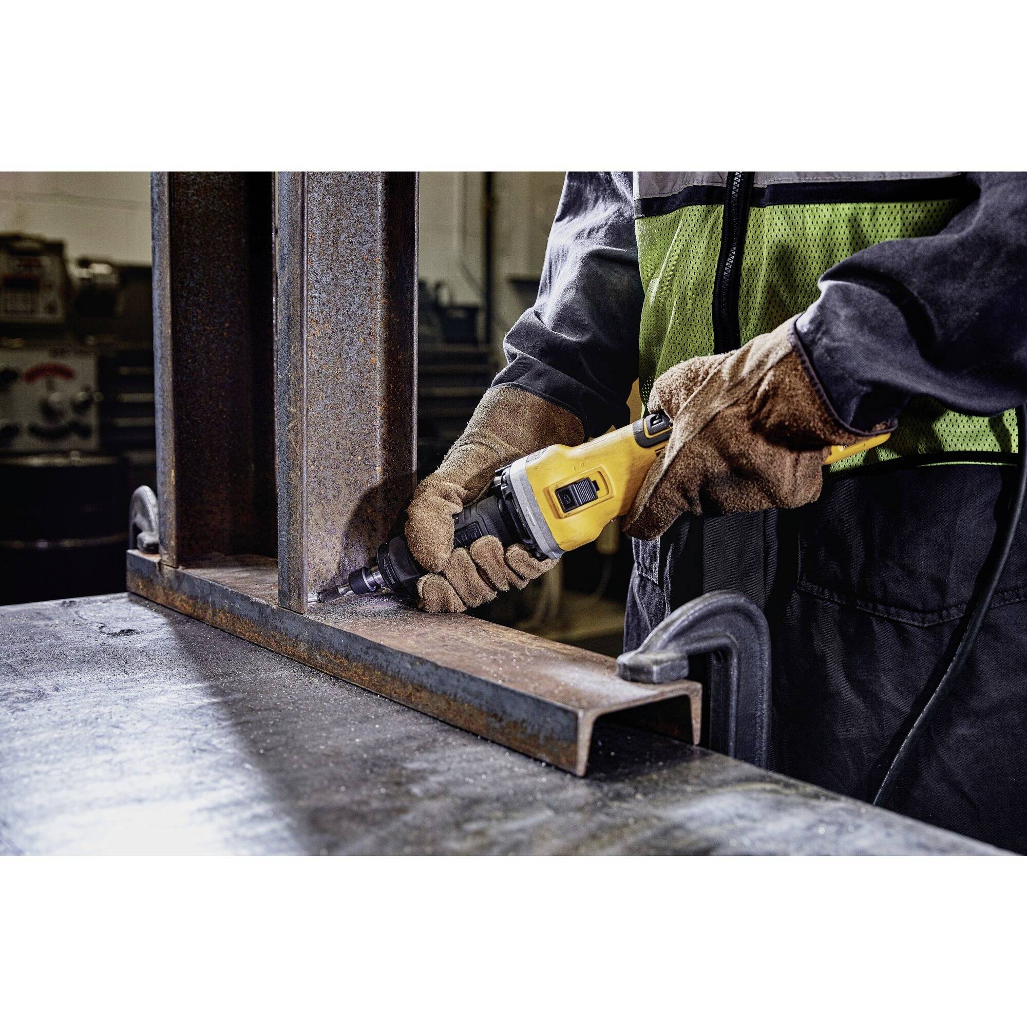 Une personne travaille avec une meuleuse d'angle jaune sur un support métallique dans un atelier. Elle porte une combinaison de protection et des gants.