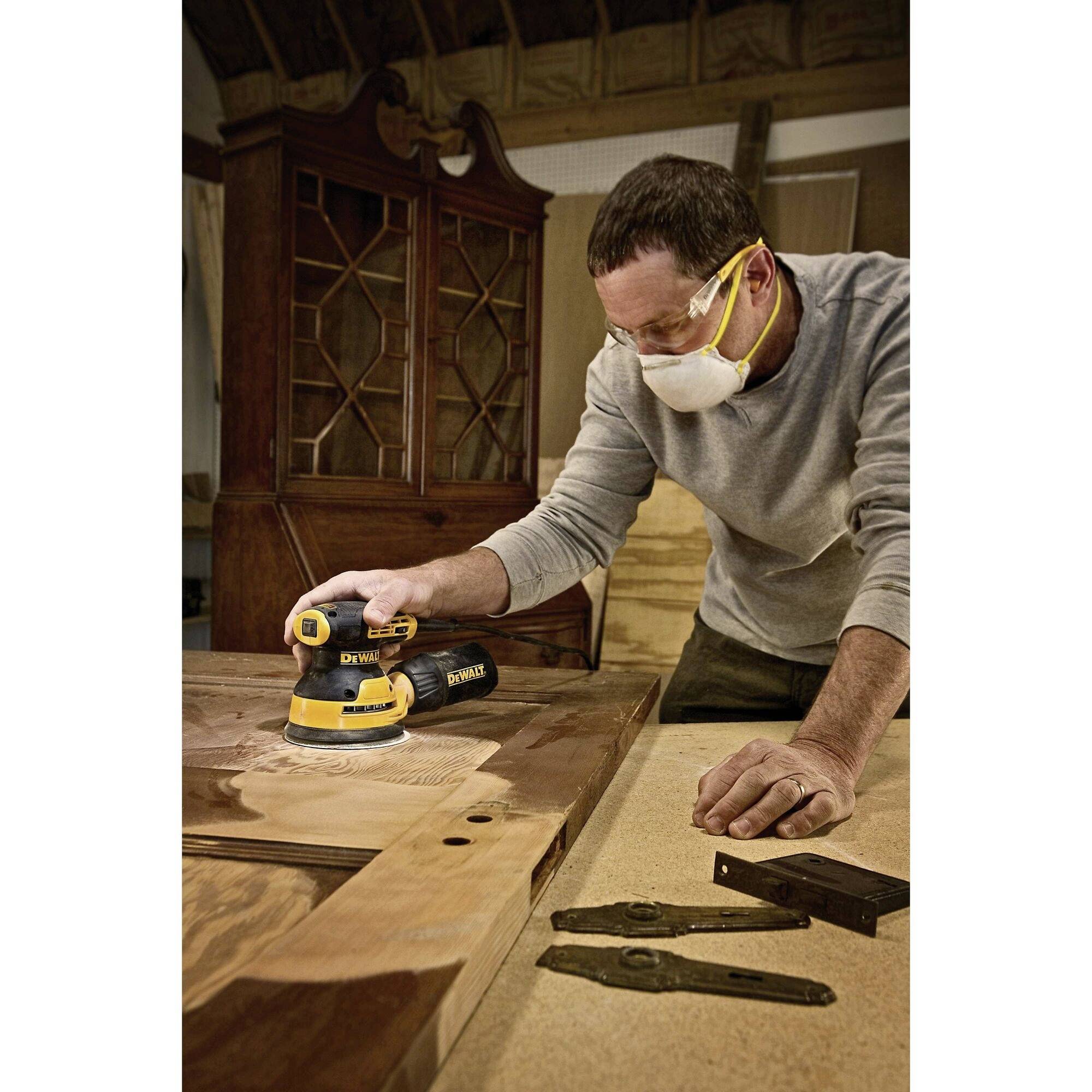 Une personne ponçe une planche de bois à l'aide d'un outil de ponçage électrique, portant un masque de protection et des lunettes de sécurité.