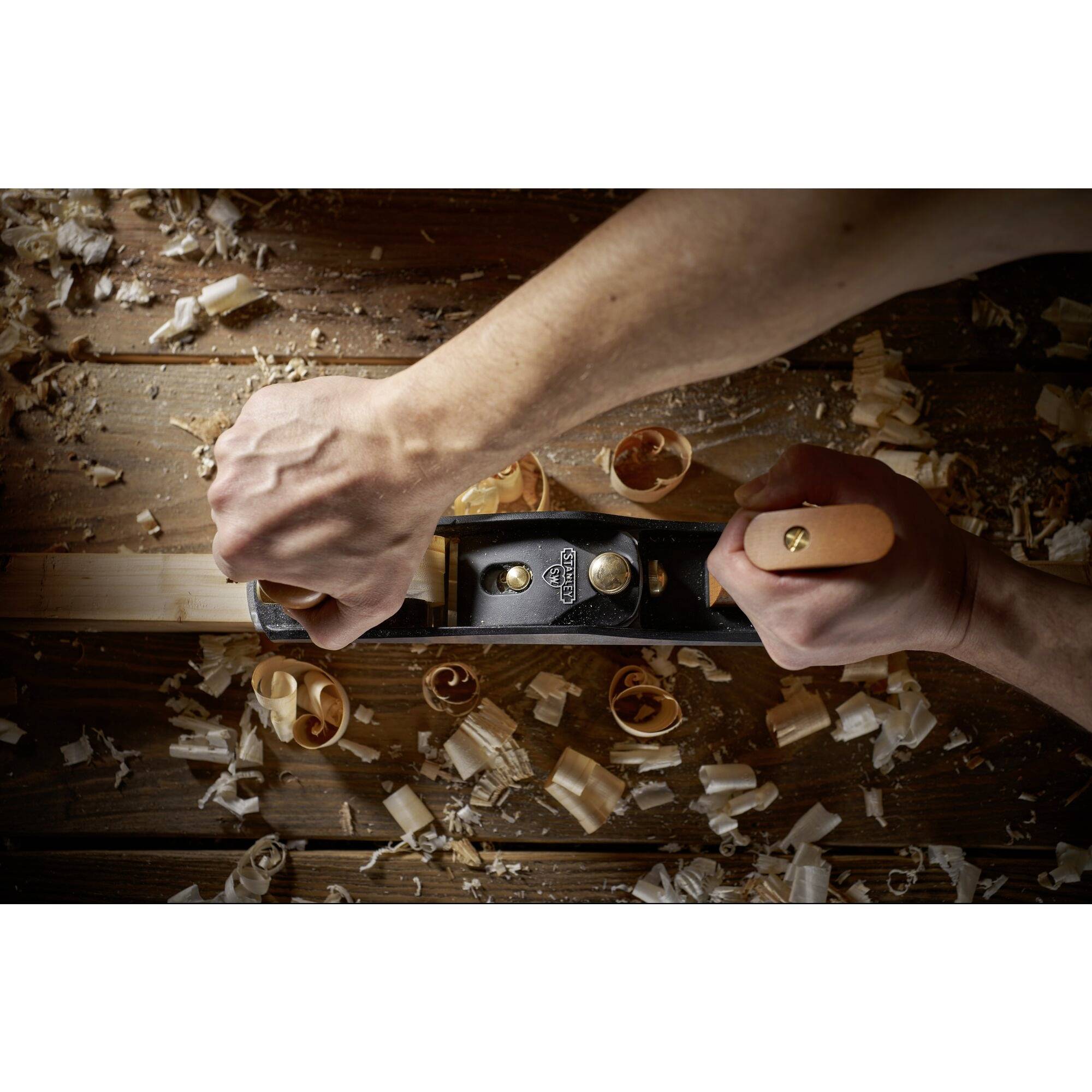 Deux mains utilisent un rabot pour lisser du bois sur une table. Des copeaux de bois sont éparpillés, ce qui indique un travail artisanal.