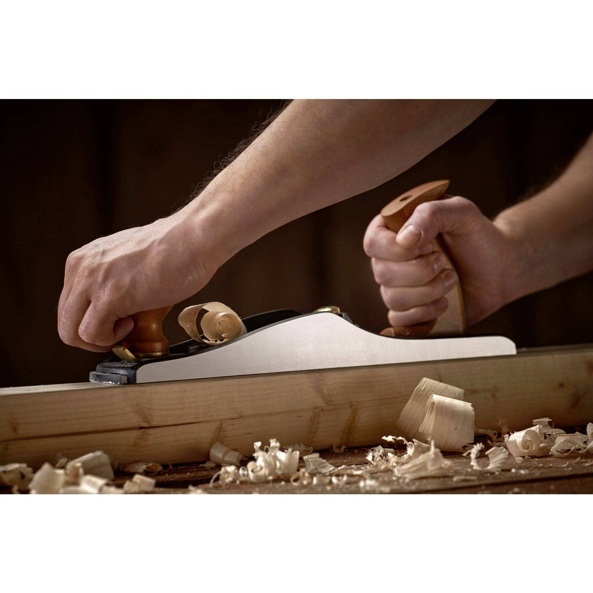 Travail du bois : Une personne rabote une planche de bois à l'aide d'un rabot à main, entourée de copeaux de bois.