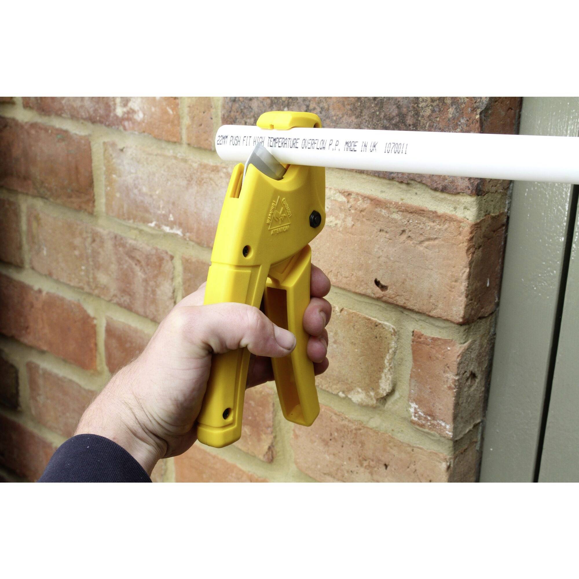 Une main tient une pince coupe-tube jaune qui coupe un tuyau blanc en PVC devant un mur de briques.