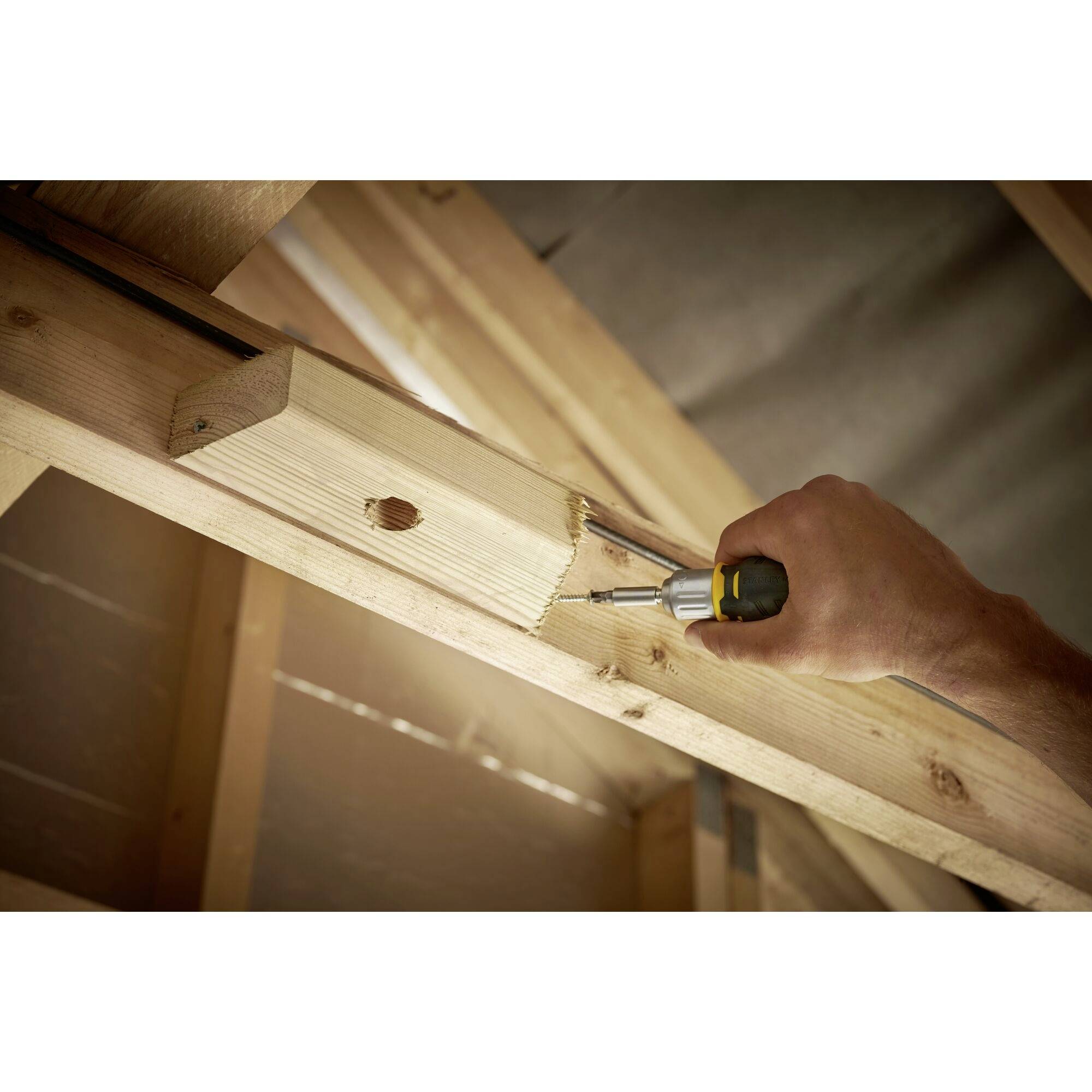 Un artisan scie une planche de bois sur un chevron pour l'ajuster. L'arrière-plan montre la structure en bois non couverte d'un grenier.