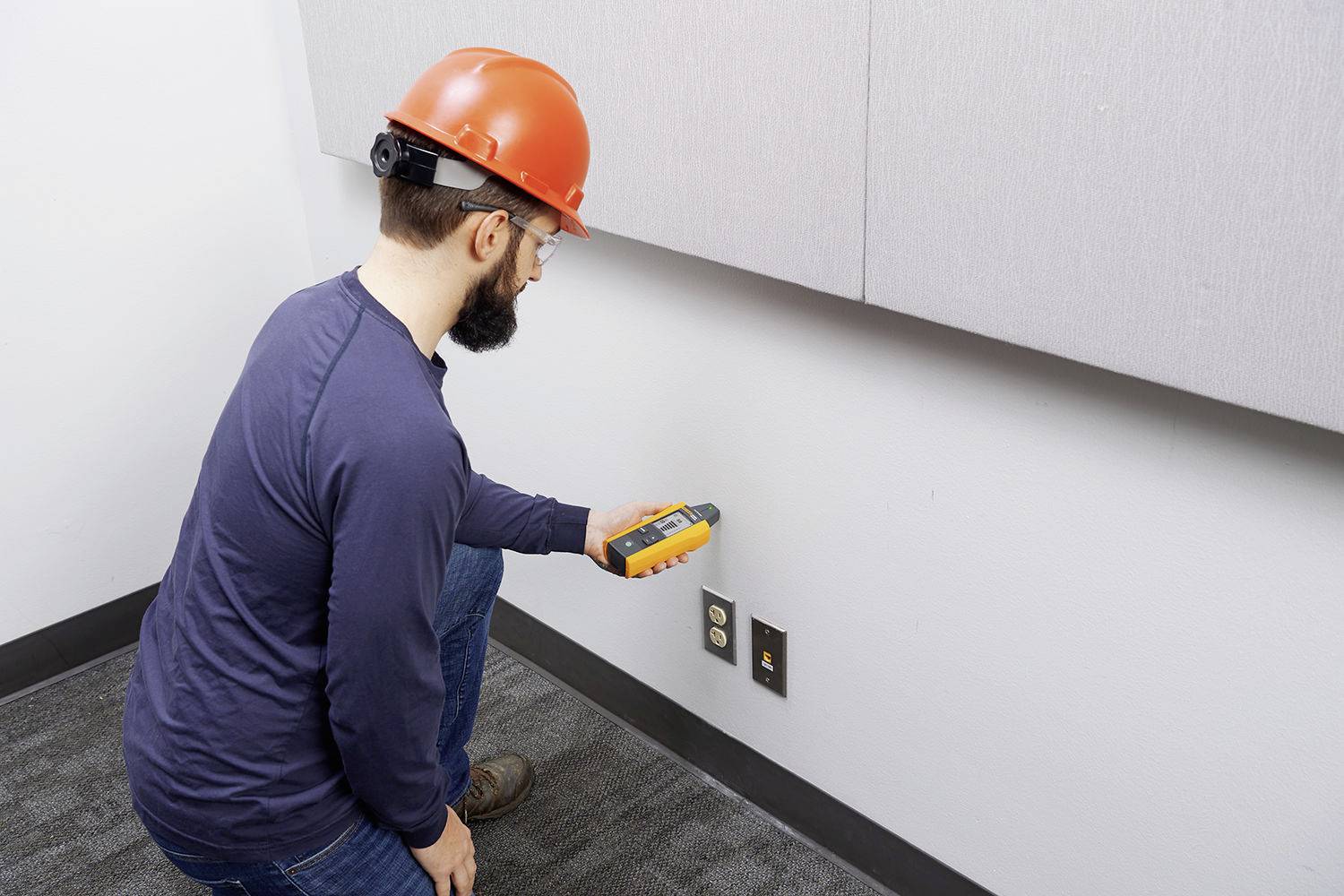 Un homme portant un casque de protection vérifie une prise électrique sur un mur à l'aide d'un appareil de mesure.