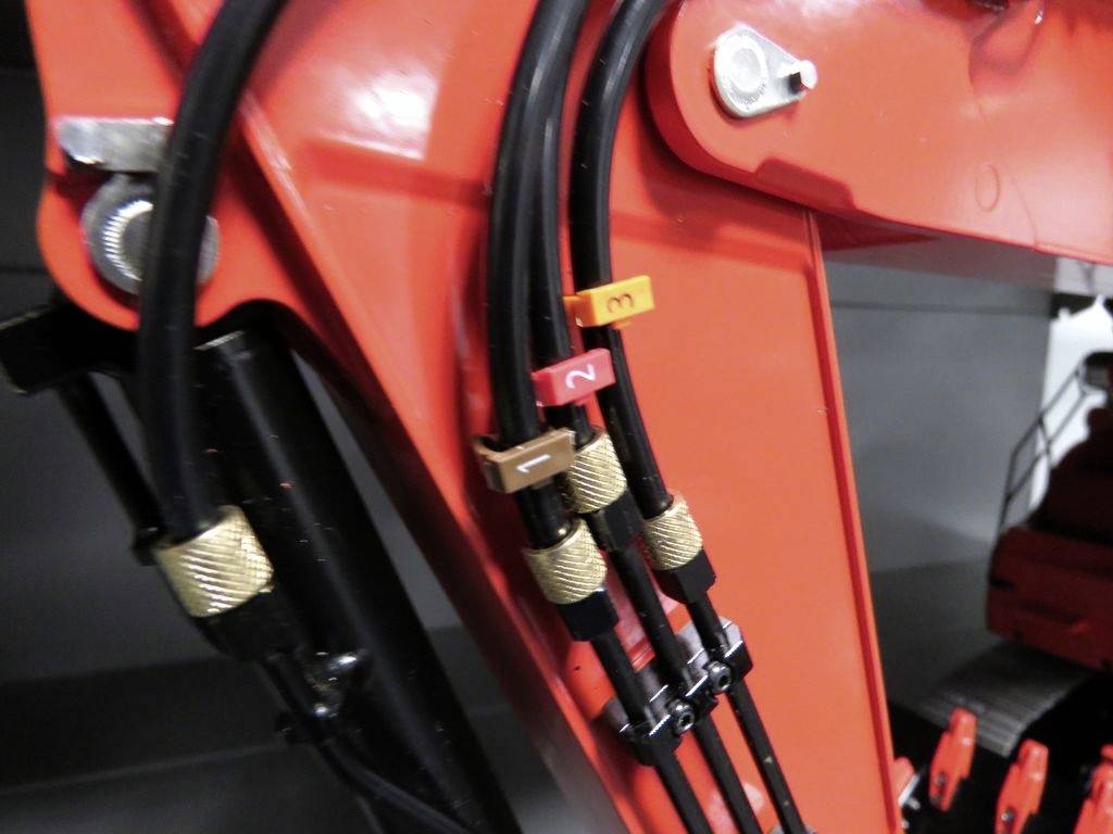 Gros plan sur des tuyaux hydrauliques sur une partie d'engin de chantier rouge, étiquetés avec des clips en plastique colorés indiquant les positions des tuyaux.