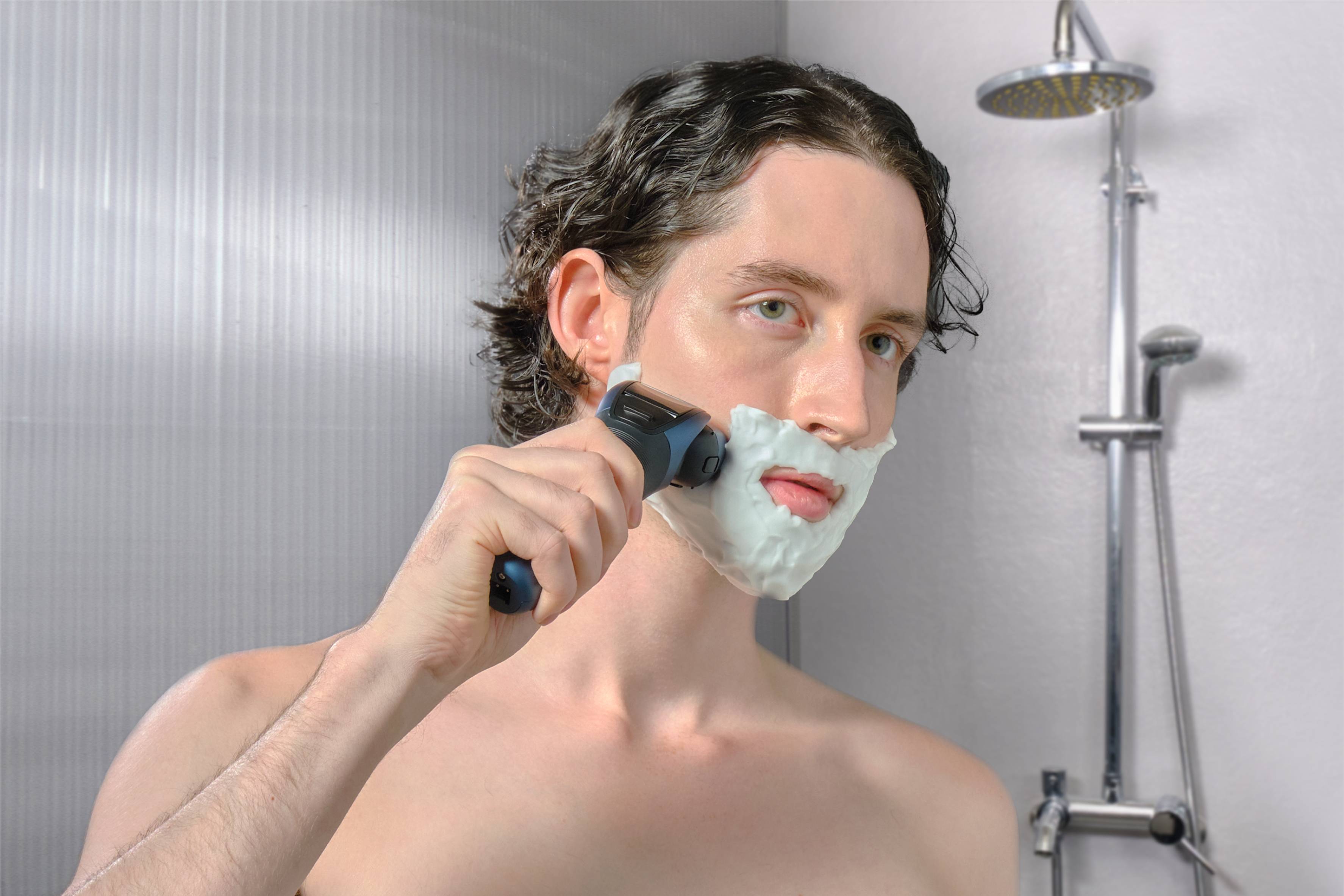Un homme se rase avec un rasoir électrique dans la salle de bain. Il a de la mousse à raser sur le visage et se trouve debout devant une douche.