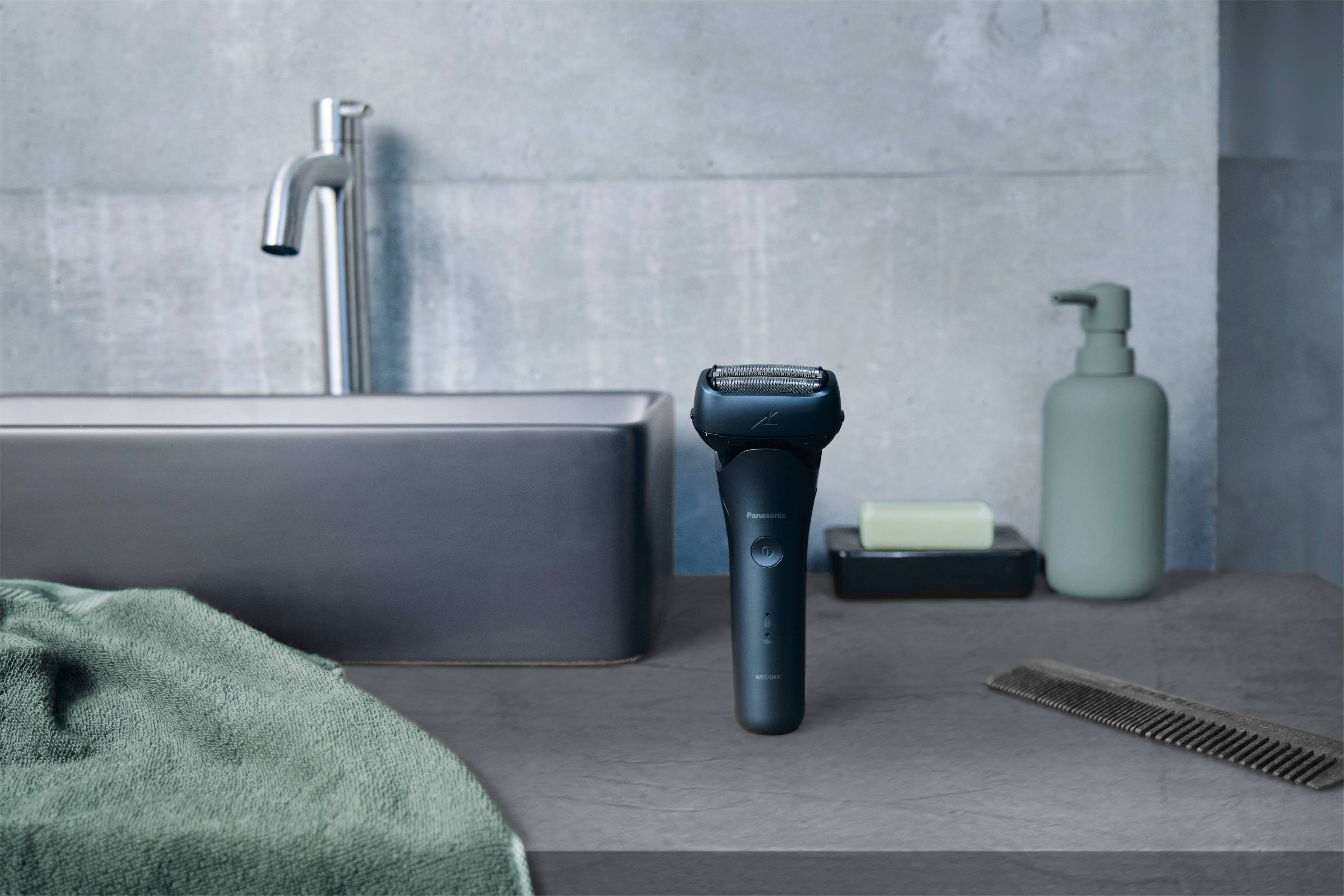 Rasoir électrique sur un comptoir gris de salle de bain, à côté du lavabo, du distributeur de savon et du peigne.