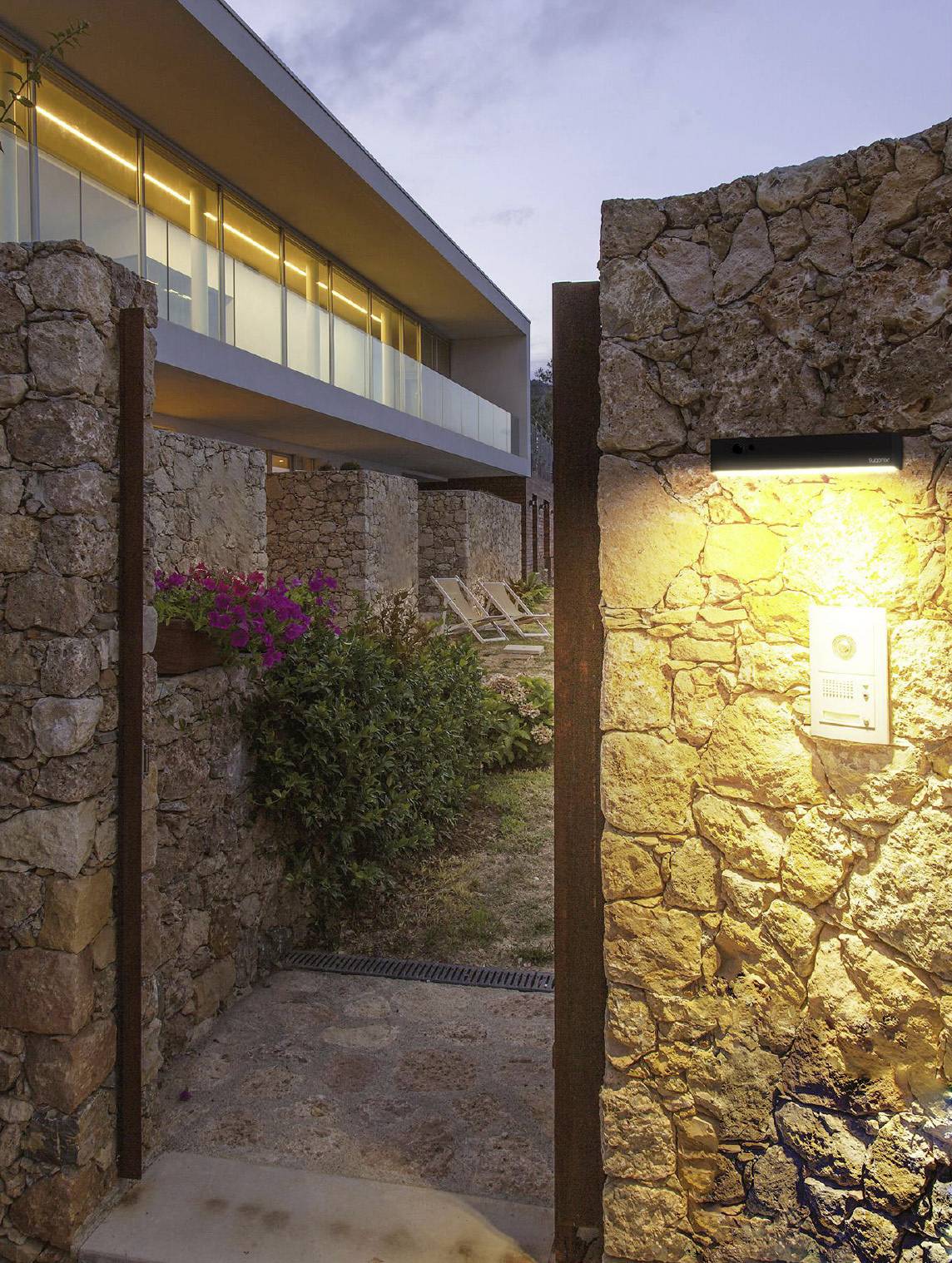 Mur de pierre avec un portail éclairé ouvrant la voie vers un bâtiment moderne à façade vitrée. Fleurs et espaces de repos dans le jardin.