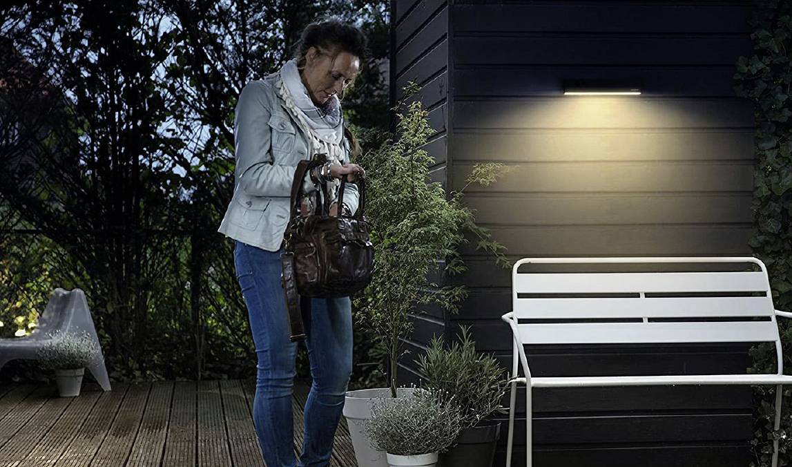 Une personne se tient la nuit devant un banc éclairé sur une terrasse et regarde dans un sac à main. Des plantes sont en arrière-plan.