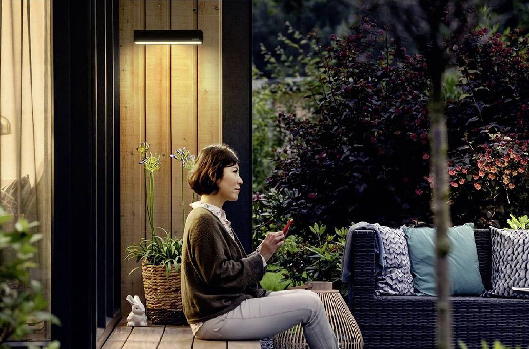 Une femme est assise dehors sur une terrasse, entourée de plantes, et utilise un smartphone. C'est le soir et une lumière de bougie éclaire l'espace.