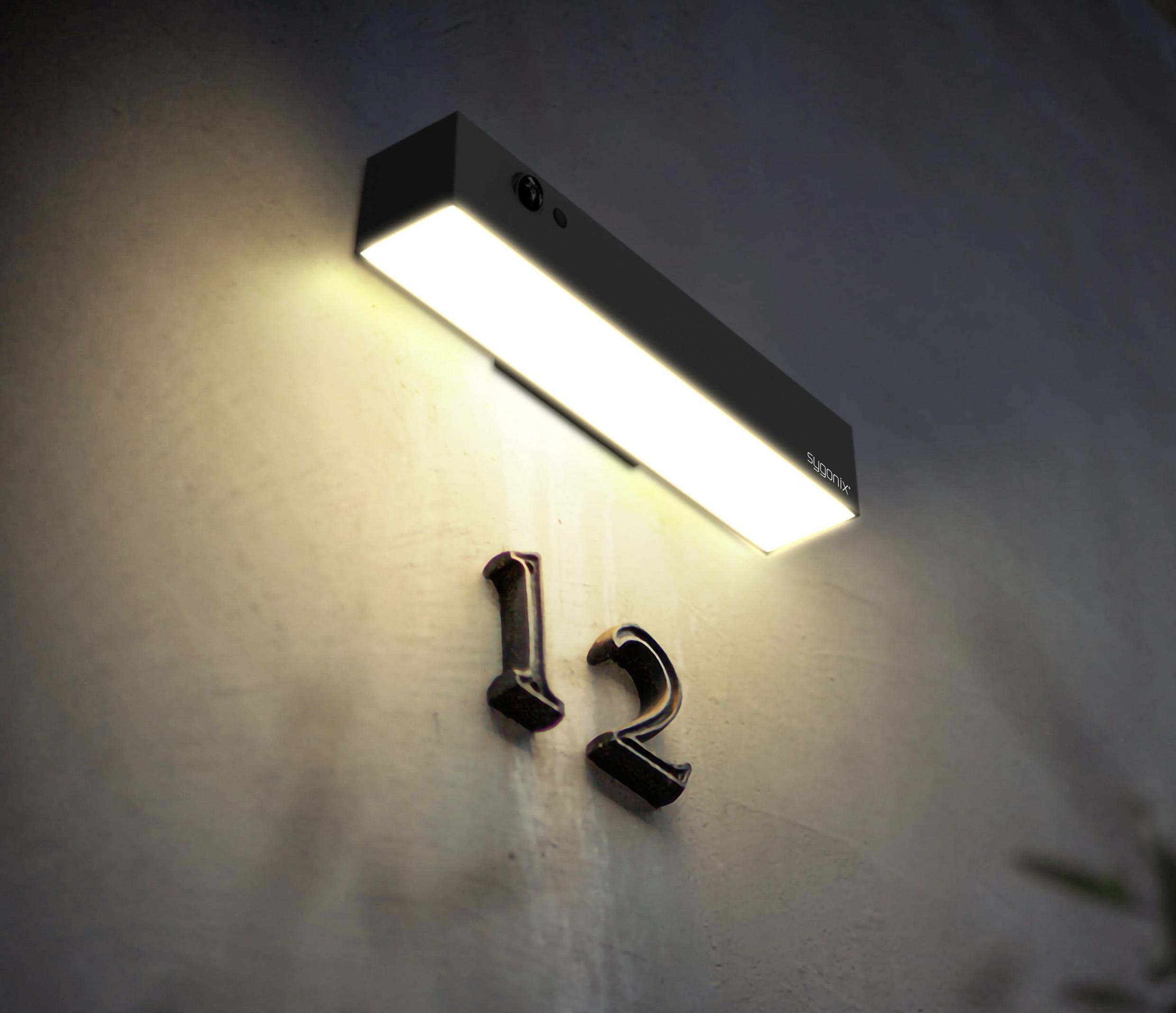 Un luminaire extérieur éclaire le numéro de maison '12' sur un mur au crépuscule.