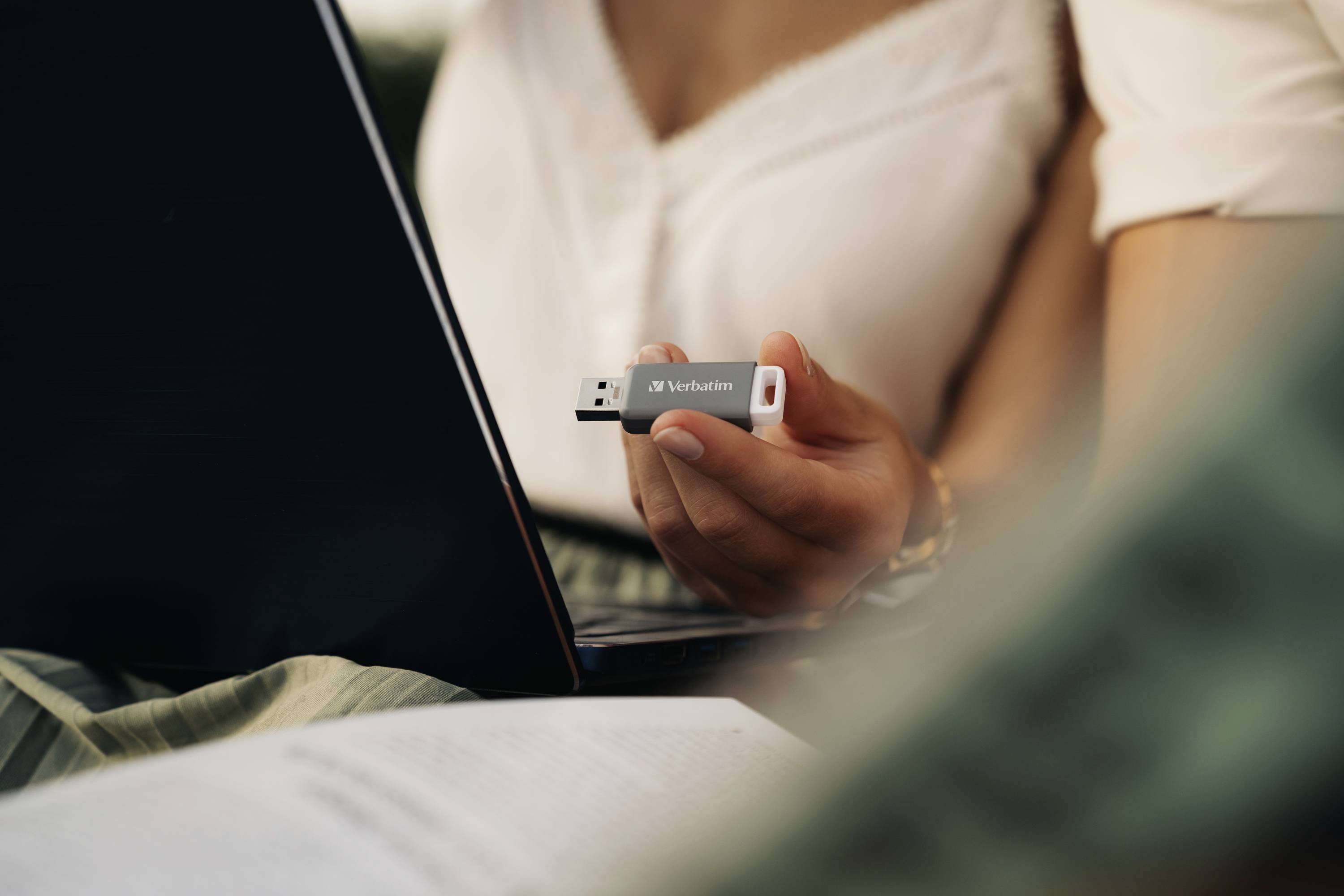 Une personne tient une clé USB et l'insère dans un ordinateur portable. Un livre ouvert est au premier plan.