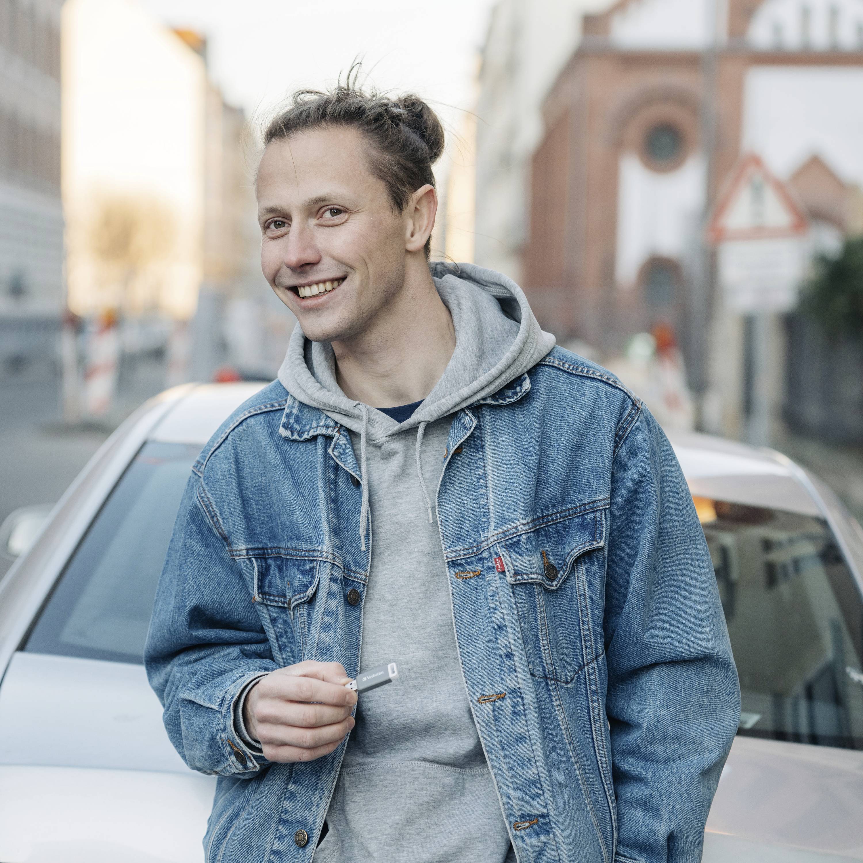 Une personne portant une veste en jean et un sweat à capuche gris se tient souriante devant une voiture sur une rue urbaine.