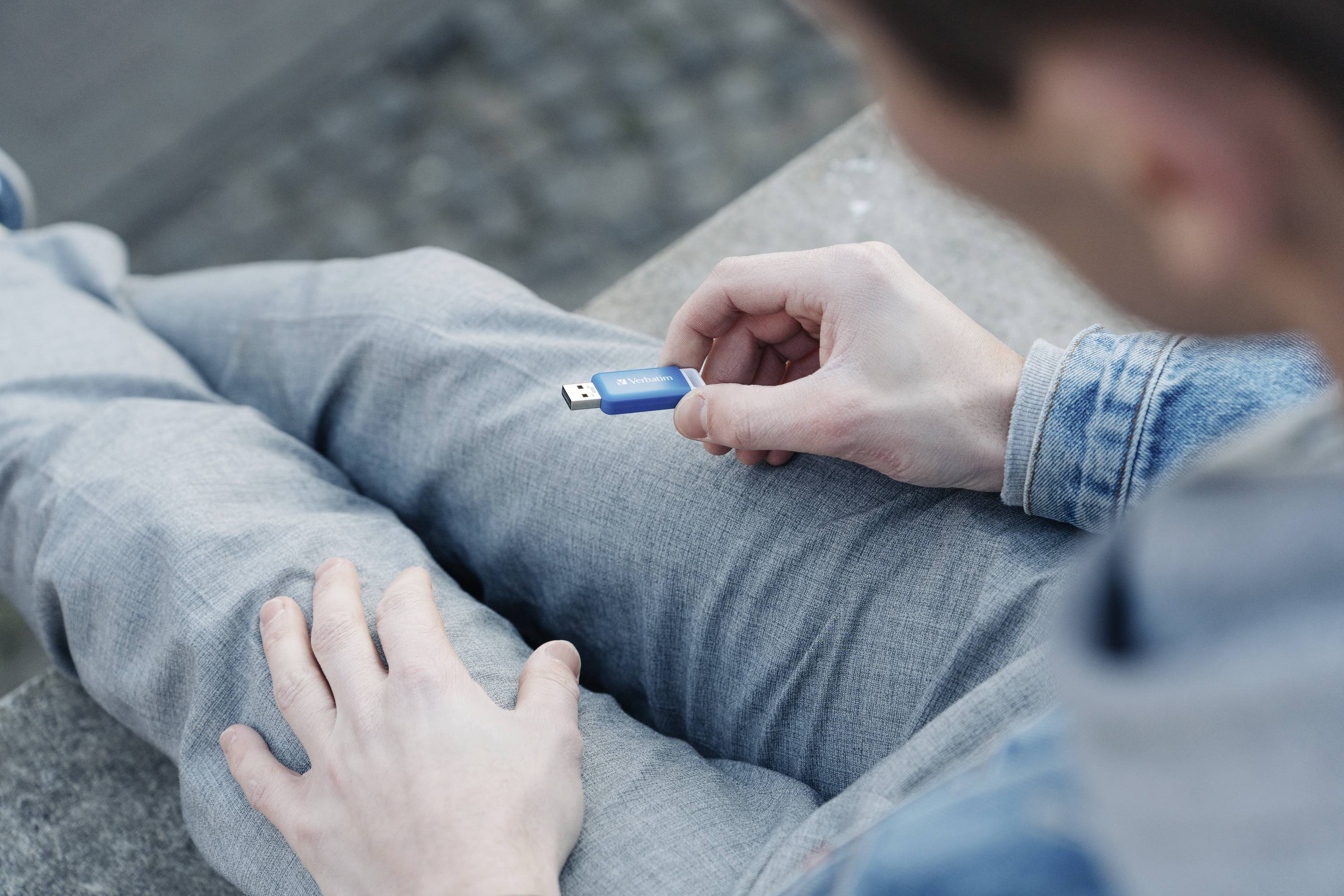Une personne tient une clé USB bleue dans sa main et est assise sur un pantalon gris. La scène est vue de dessus.