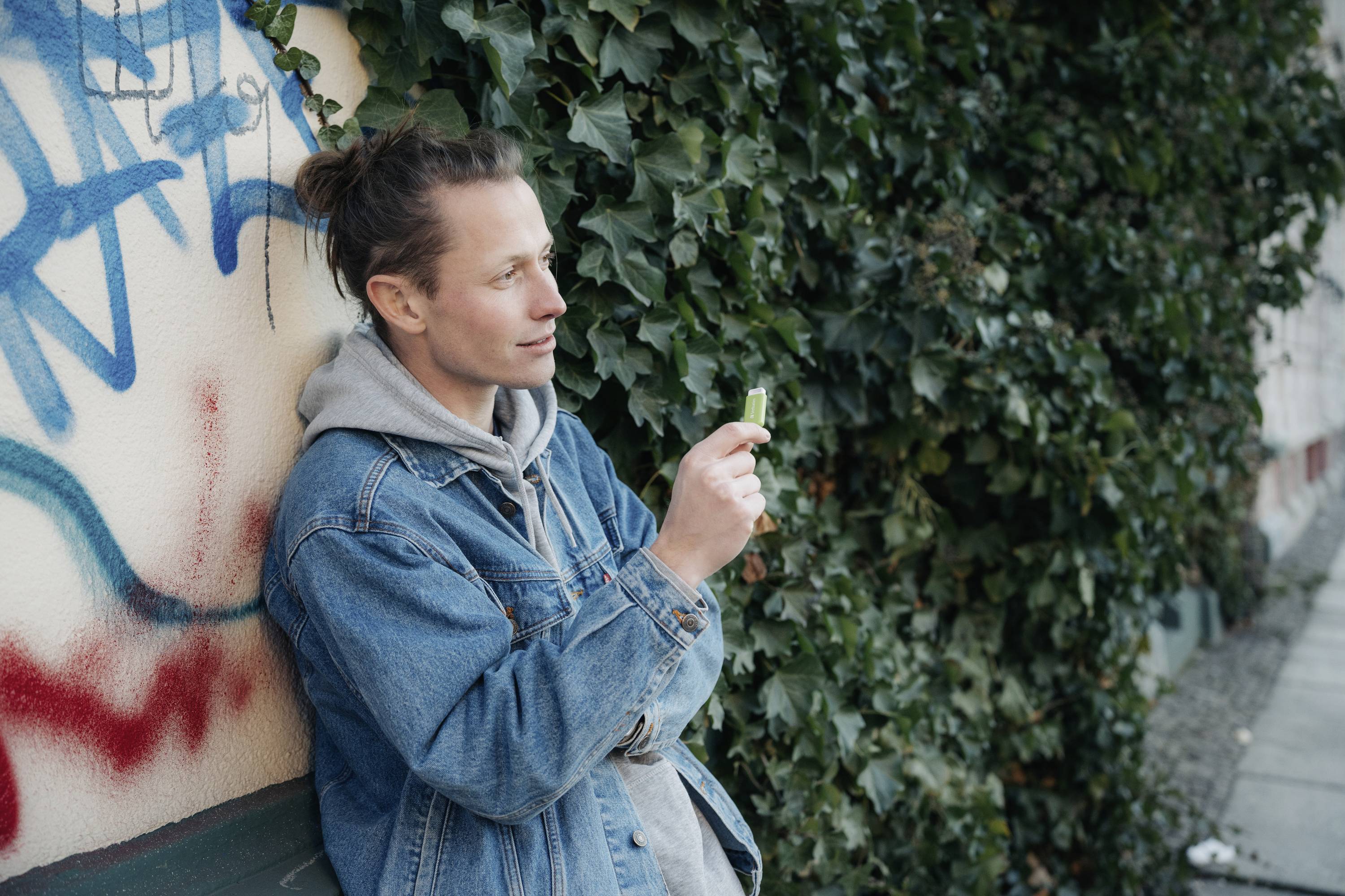 Une personne portant une veste en jean se tient contre un mur couvert de lierre, tenant un petit objet et paraissant pensive.