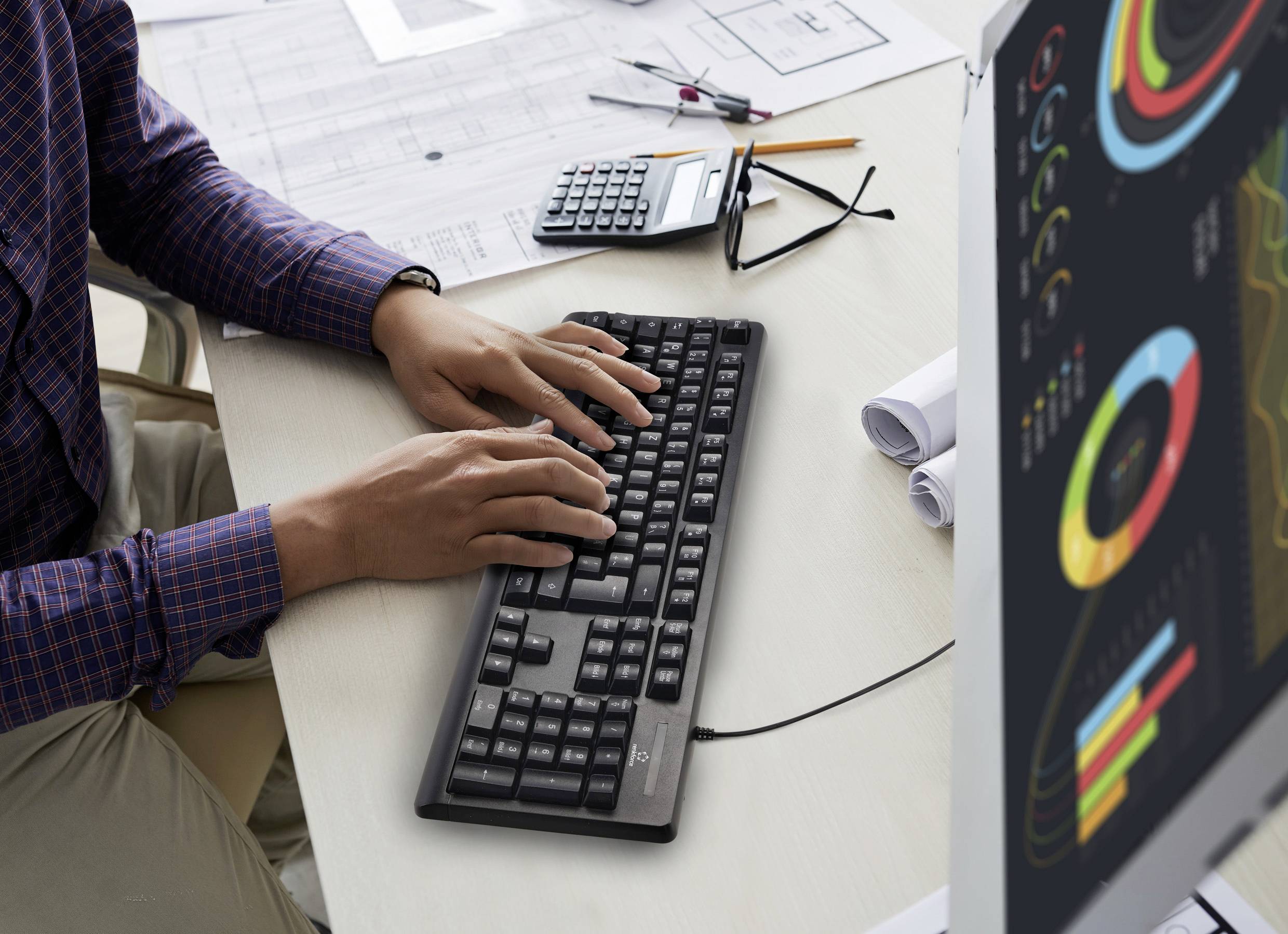 Une personne tape sur un clavier, installée à un bureau avec des diagrammes à l'écran, des documents et une calculatrice.