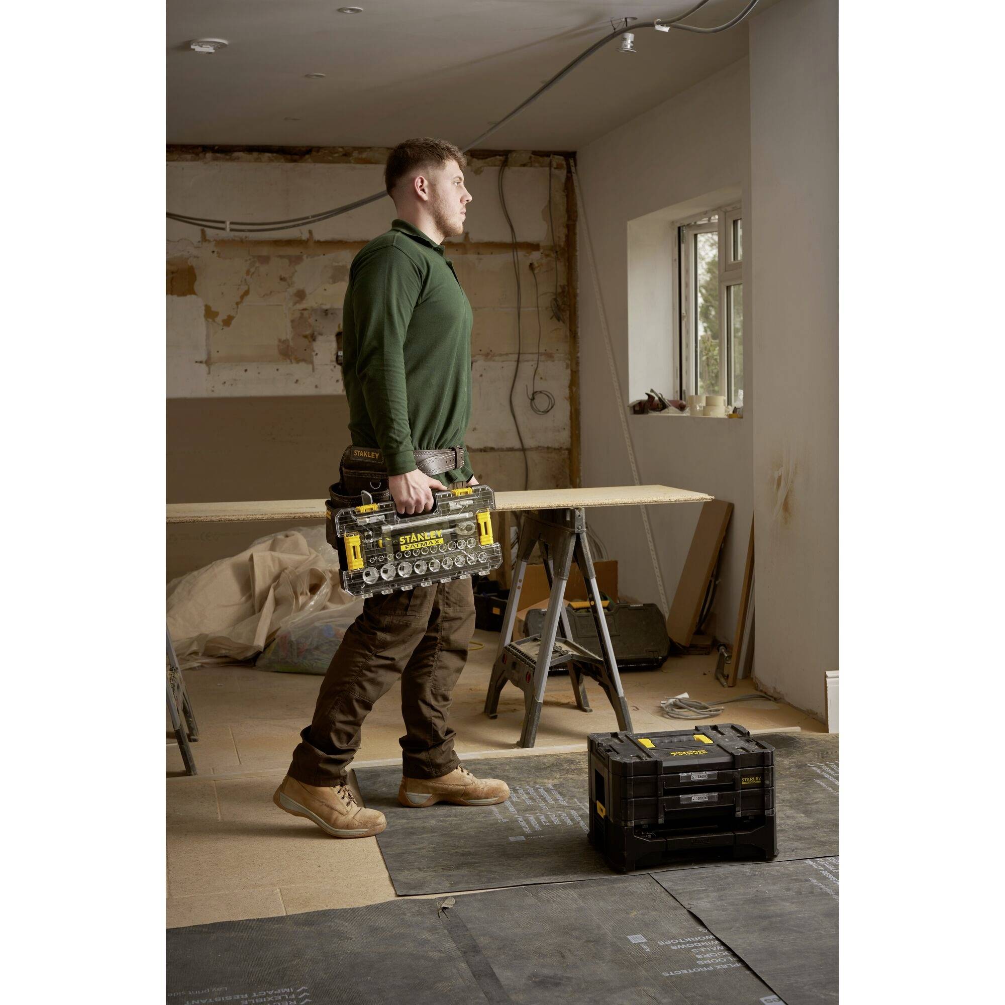 Un homme transporte une boîte à outils dans un atelier en construction avec des fenêtres. Des tréteaux et des outils sont visibles en arrière-plan.