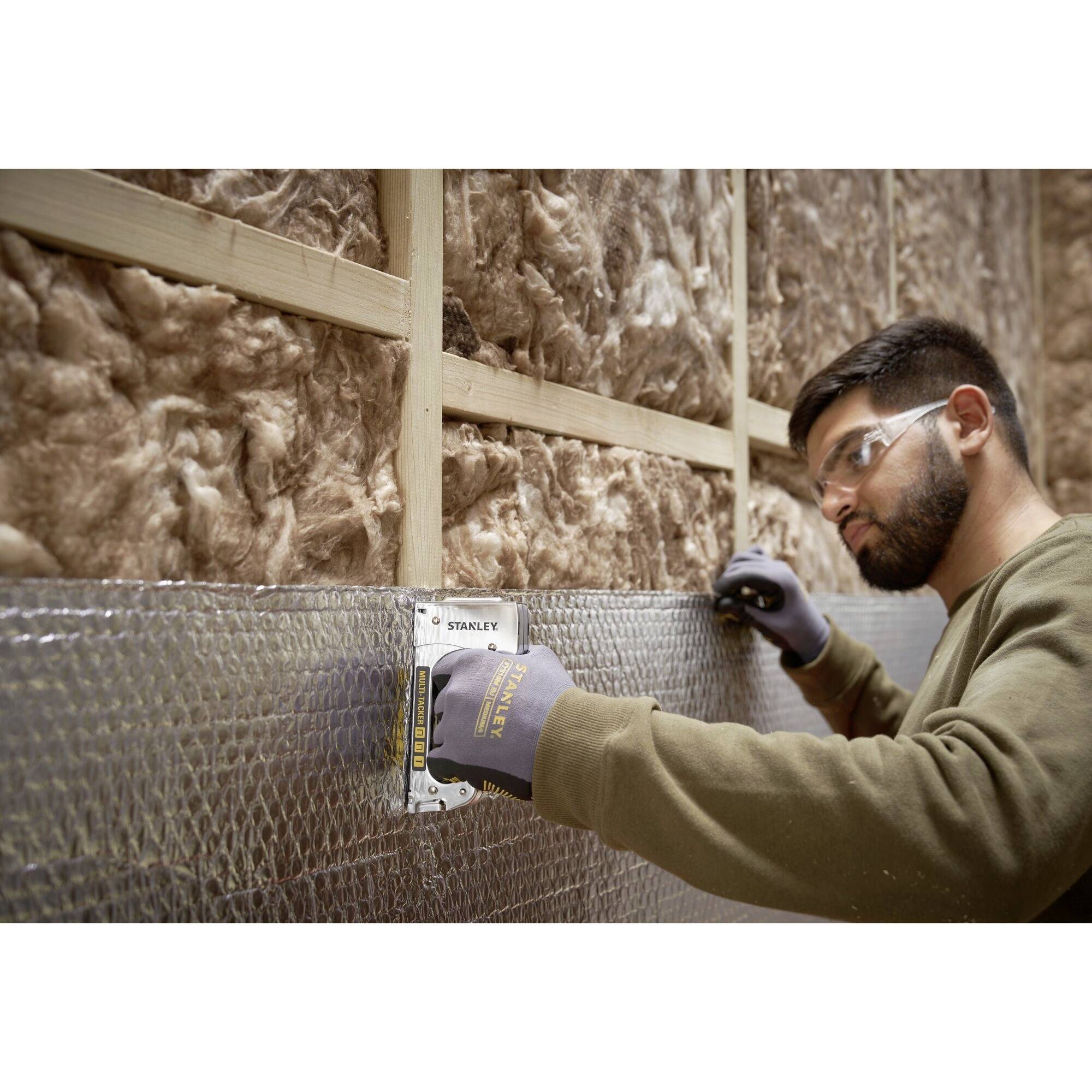 Un homme portant des lunettes de protection mesure un mur isolé à l'aide d'un mètre ruban. Le mur est doté d'une couche isolante en laine de verre.