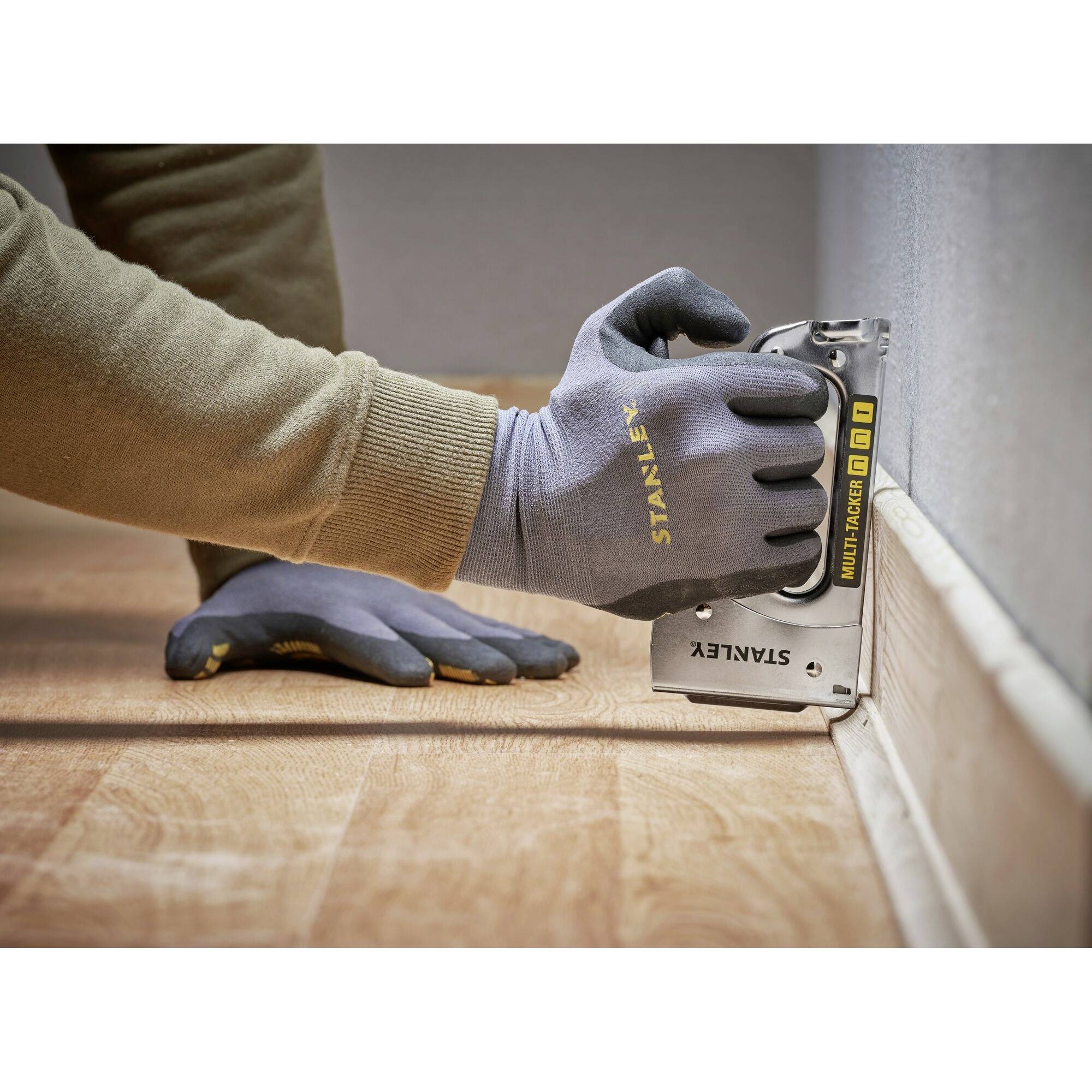 Une personne fixe des plinthes au mur à l'aide d'un pistolet à agrafes. Elle porte des gants et réalise le travail sur un sol en bois.