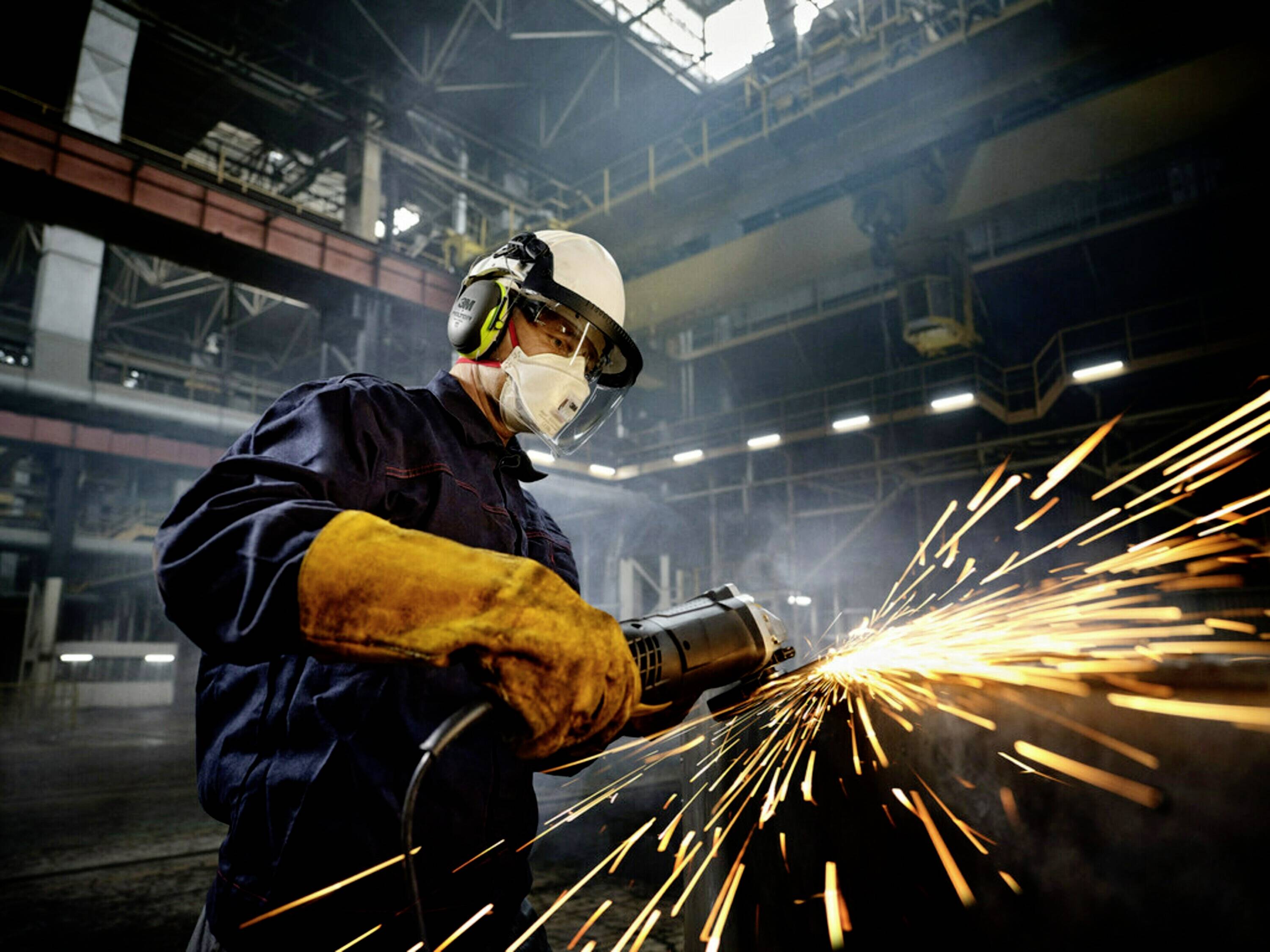 Un ouvrier en tenue de protection meule du métal avec une meuleuse d'angle dans un grand atelier industriel. Des étincelles jaillissent.
