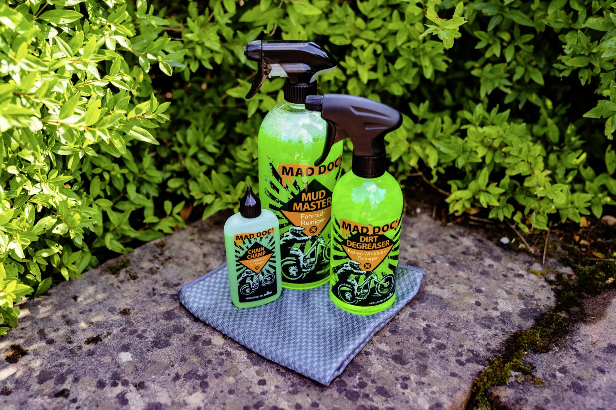 Trois produits de nettoyage dans des flacons vaporisateurs verts, posés sur un tissu gris et entourés de plantes vertes.