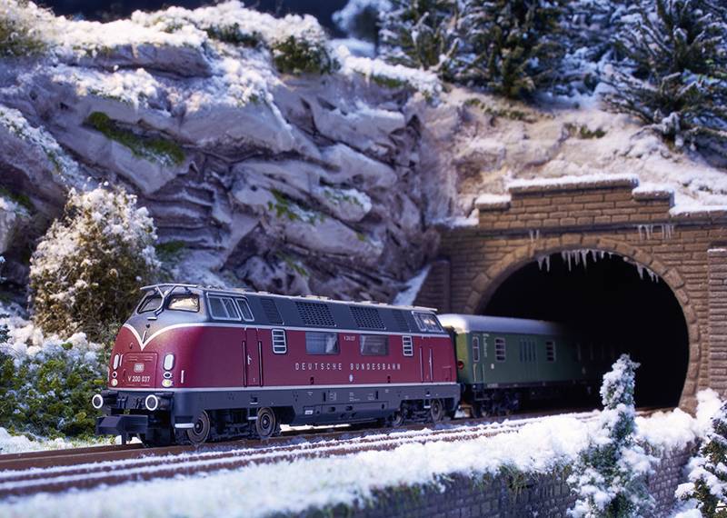 Un train miniature sort d'un tunnel dans un paysage enneigé. La locomotive est rouge, portant l'inscription 'Deutsche Bundesbahn'.