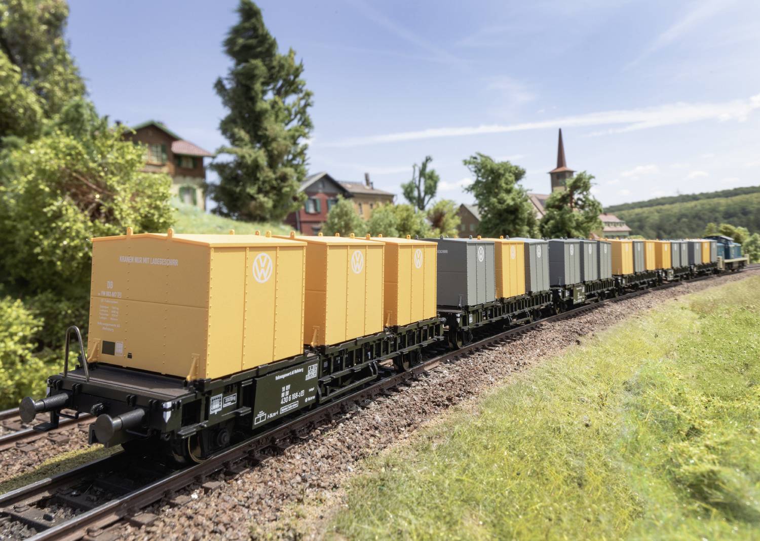 Un train miniature transportant des conteneurs de fret orange et gris traverse un paysage rural sous un ciel dégagé.