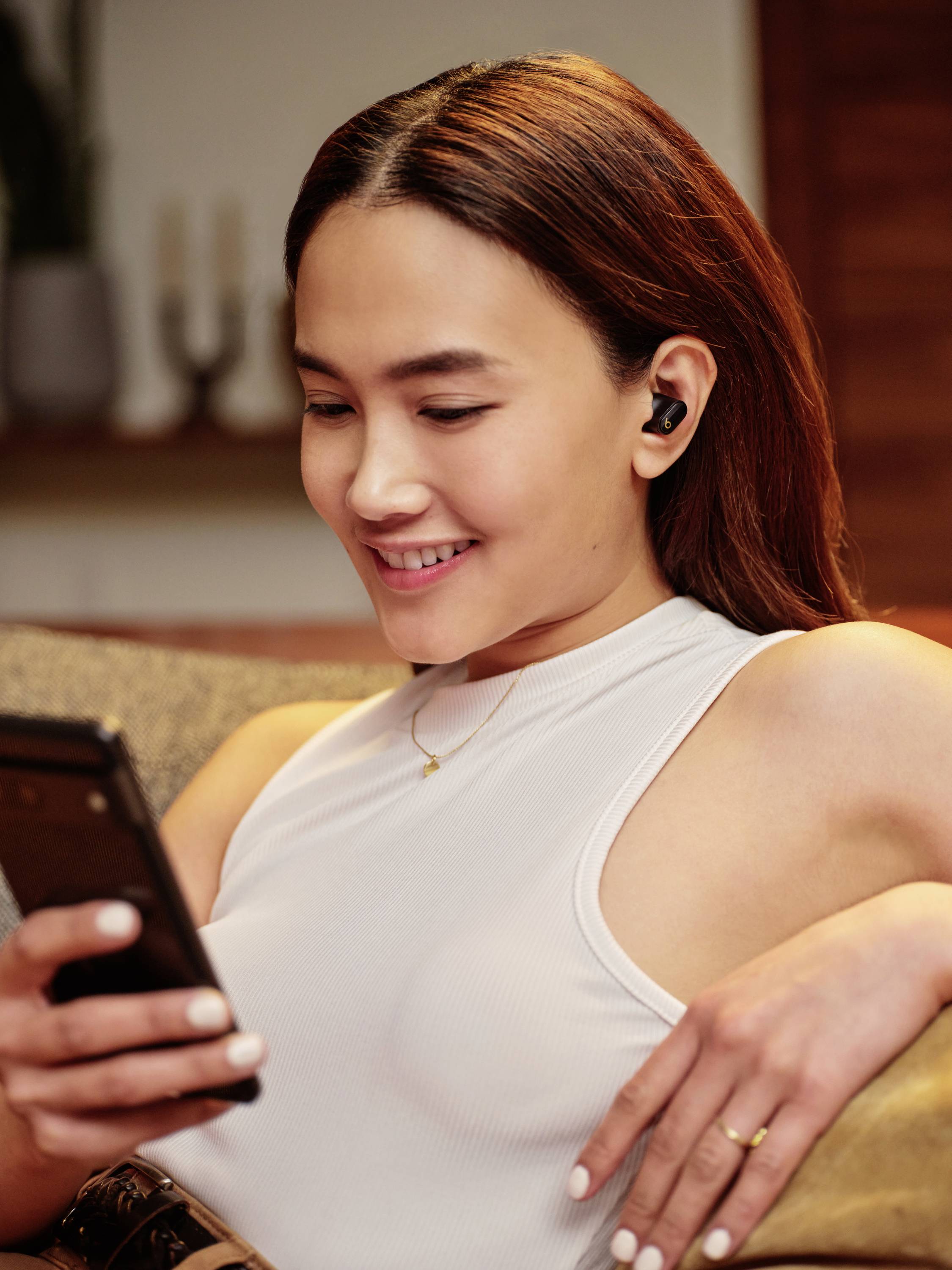 Une femme est assise sur un canapé, sourit en regardant son smartphone. Elle porte des écouteurs sans fil. Un salon est visible en arrière-plan.