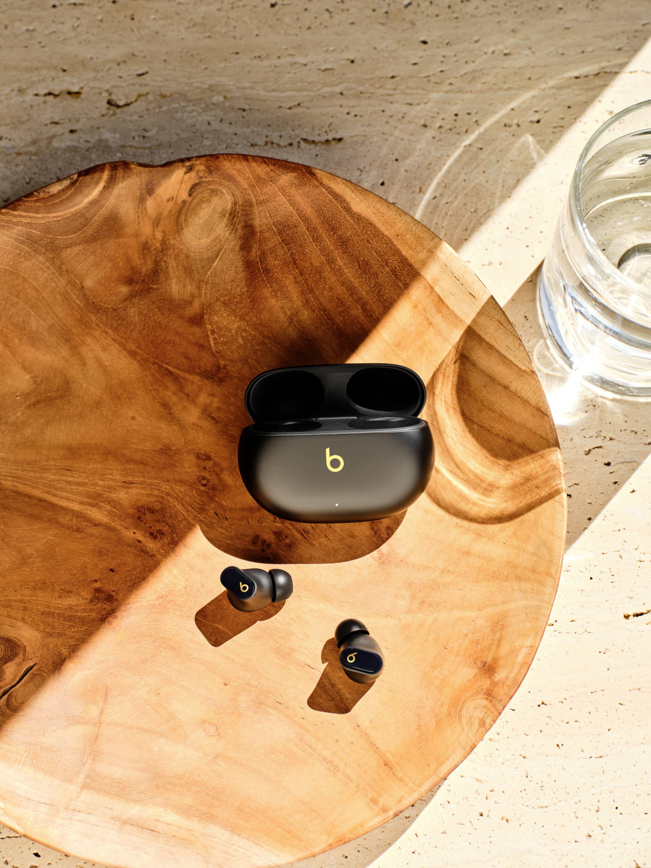 Une paire d'écouteurs sans fil et leur étui de charge ouvert sont posés sur une table ronde en bois, à côté d'un verre d'eau dans la lumière du soleil.