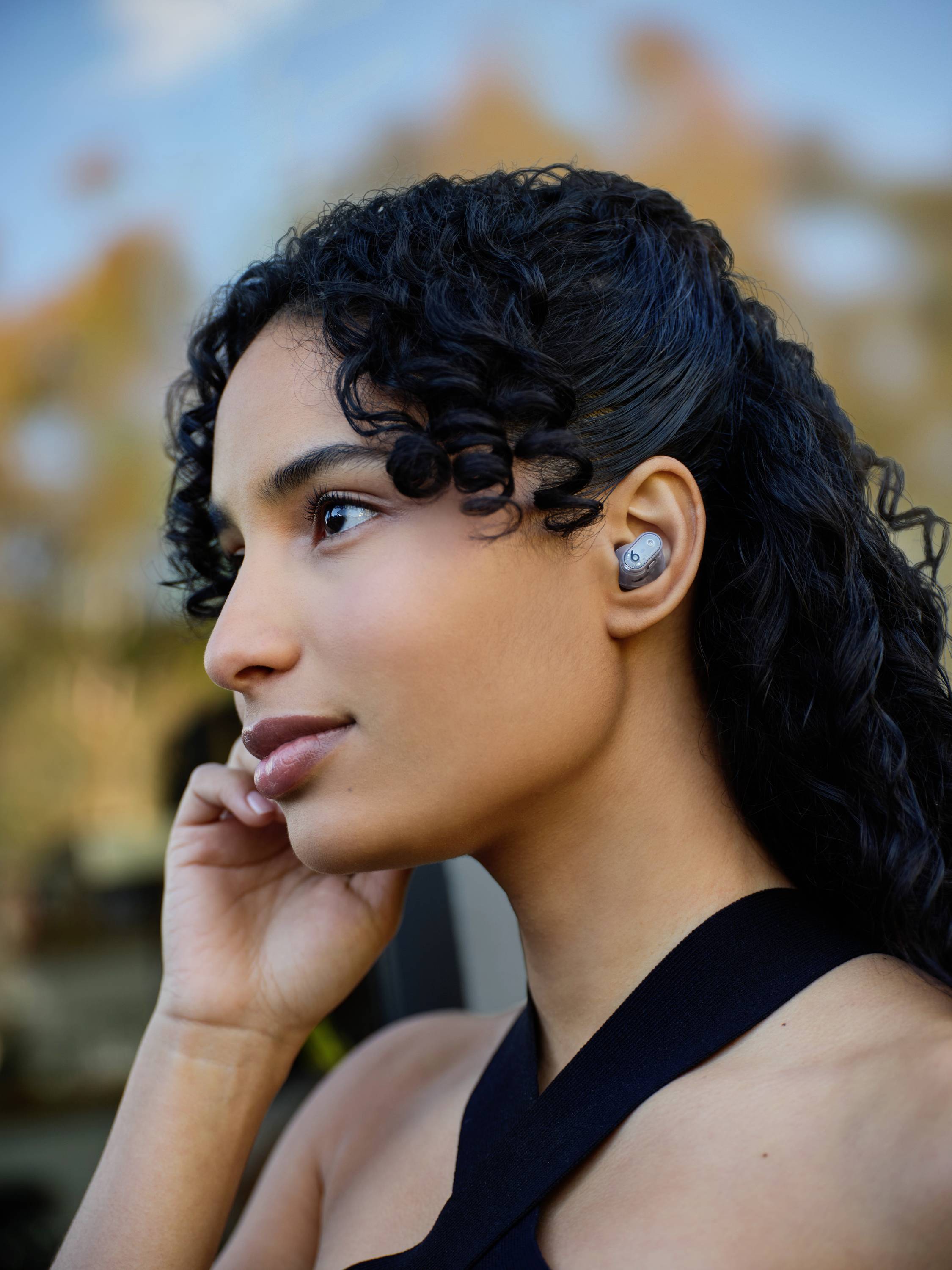 Une personne aux cheveux bouclés porte des écouteurs sans fil et regarde sur le côté, avec un arrière-plan naturel flou.