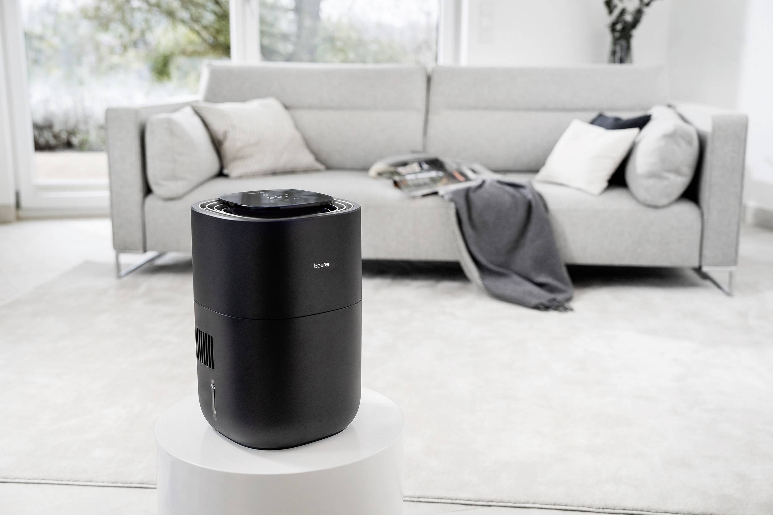 Un purificateur d'air noir est posé sur une table blanche dans un salon moderne avec un canapé gris et des coussins en arrière-plan.
