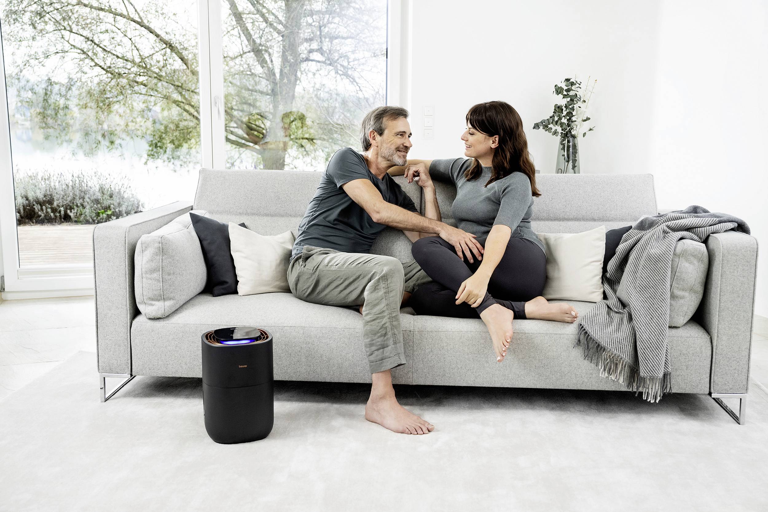 Un couple est assis sur un canapé dans un salon moderne. Ils discutent tranquillement. À côté du canapé se trouve un purificateur d'air.