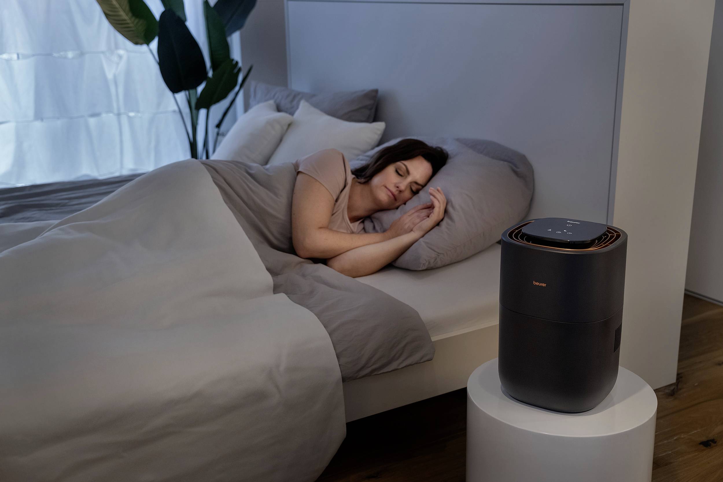 Une personne dort dans un lit dans une chambre à coucher. Un purificateur d'air est posé sur une table à côté du lit.