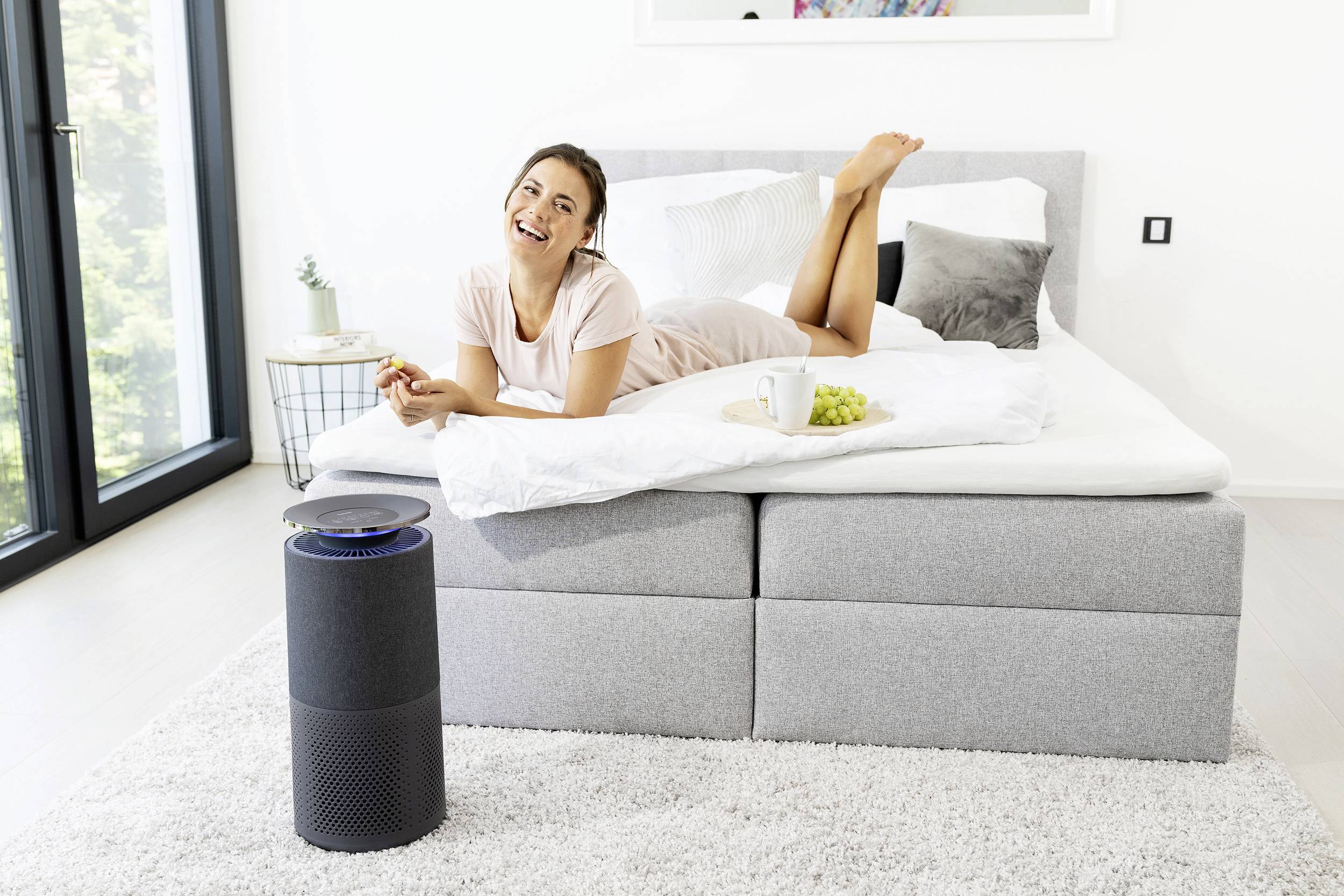 Une personne souriante est allongée de manière détendue sur un lit et mange du raisin, tandis qu'un purificateur d'air est placé à côté du lit.