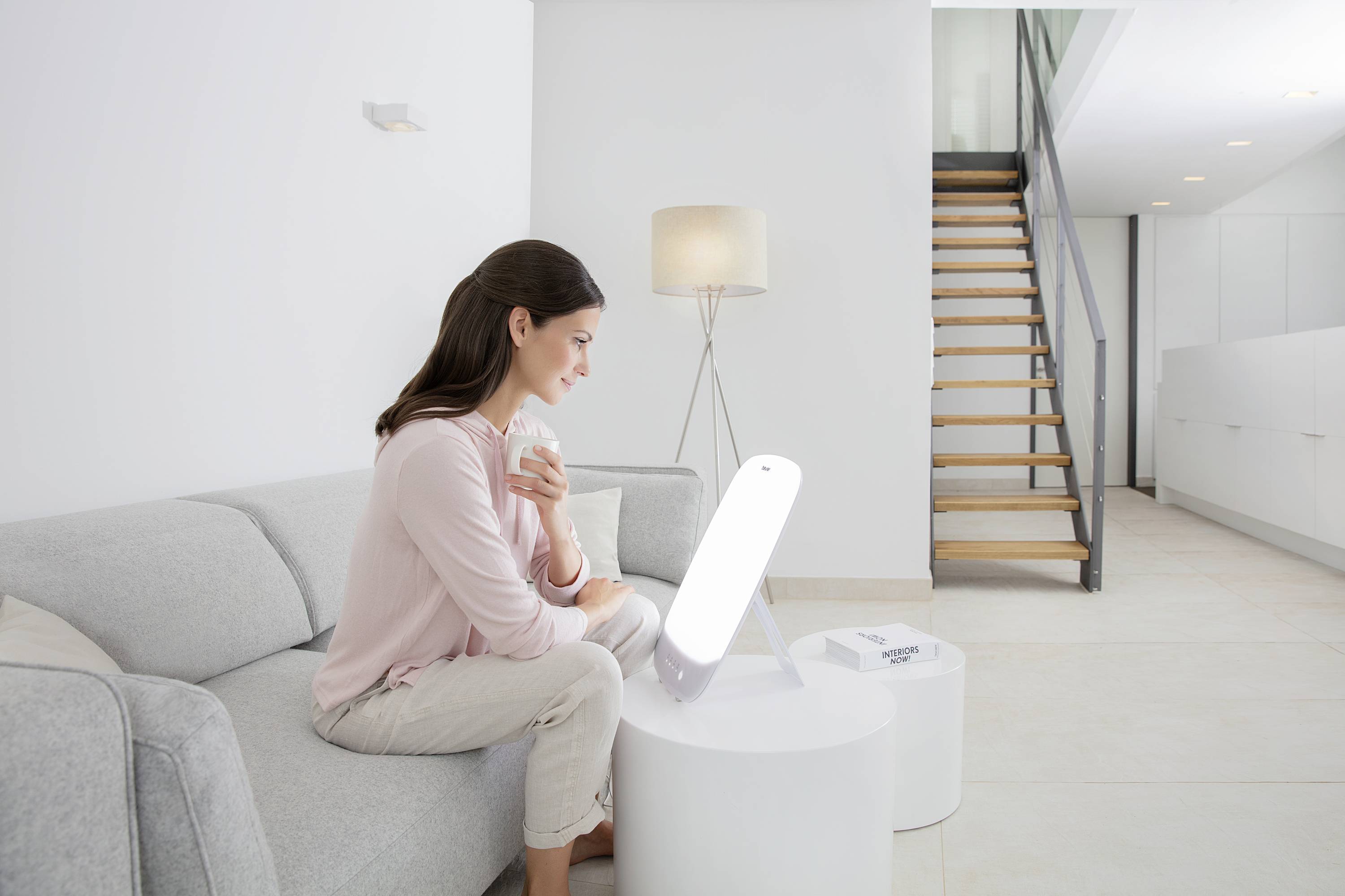 Une femme est assise sur un canapé dans une pièce lumineuse, tenant une tasse et regardant une lampe de luminothérapie posée sur une table.