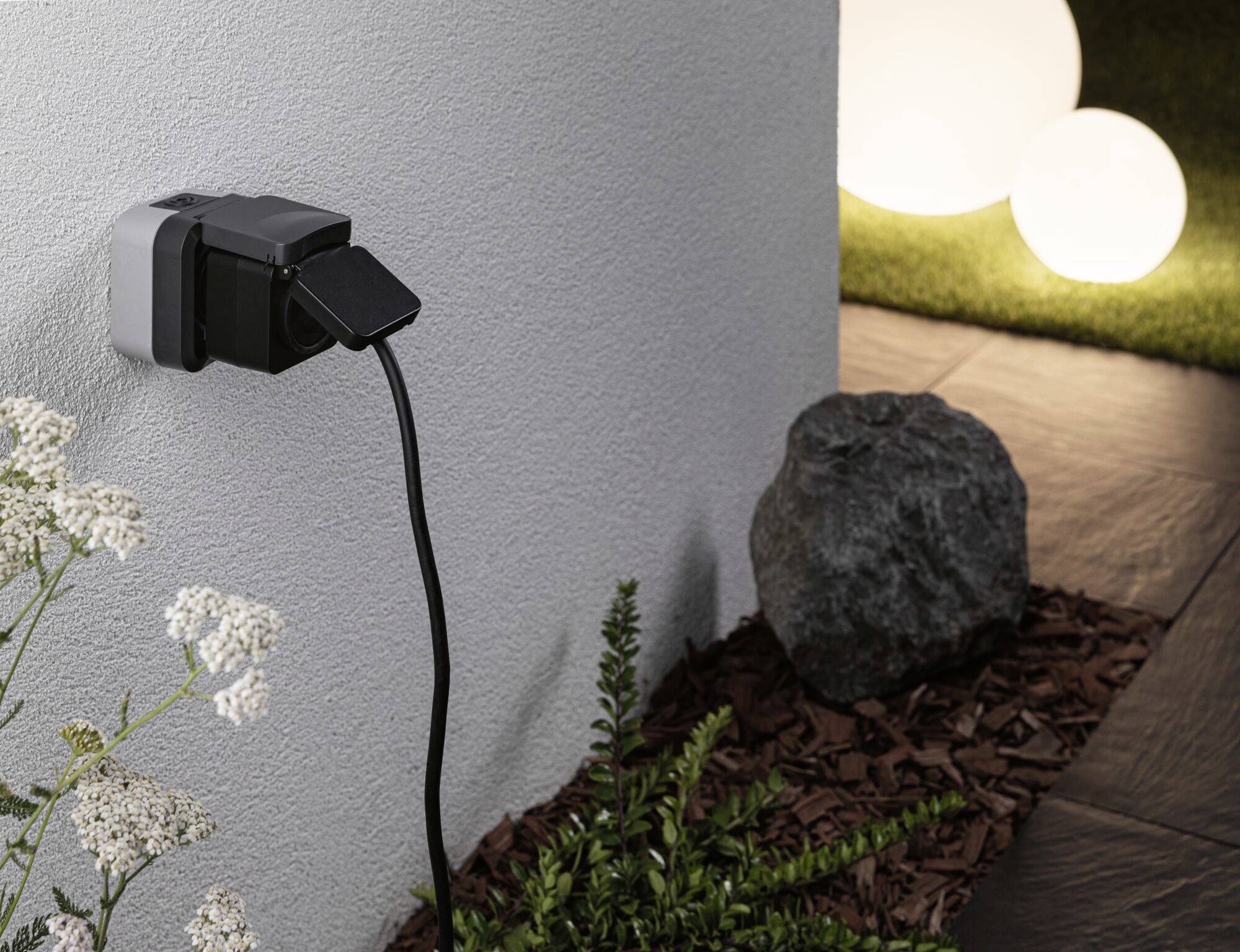 Prise électrique sur un mur extérieur avec une fiche noire branchée ; plantes et pierres au premier plan, boules lumineuses en arrière-plan.