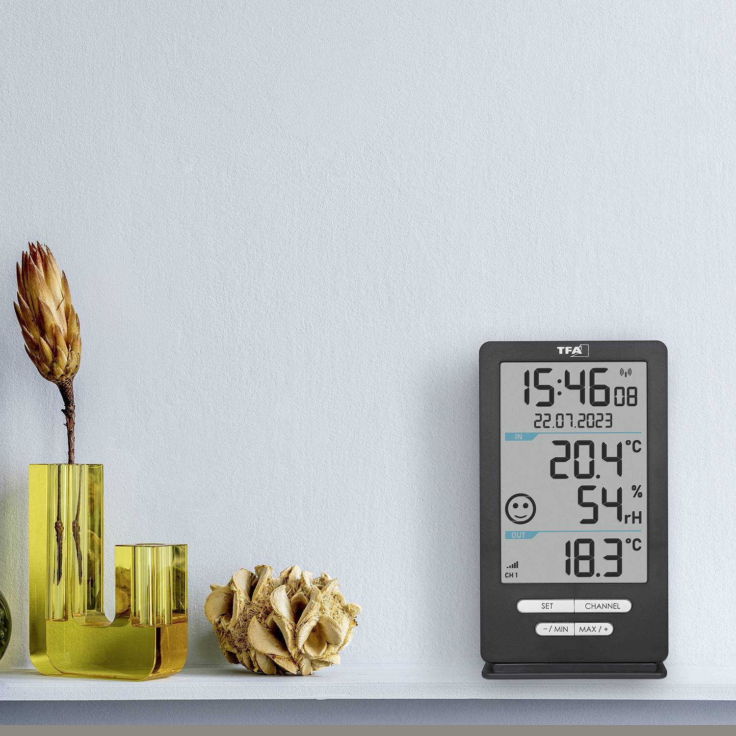 Un thermomètre numérique affiche une température de 20,4°C et une humidité de 54%. À côté se trouve un vase jaune avec une fleur séchée.