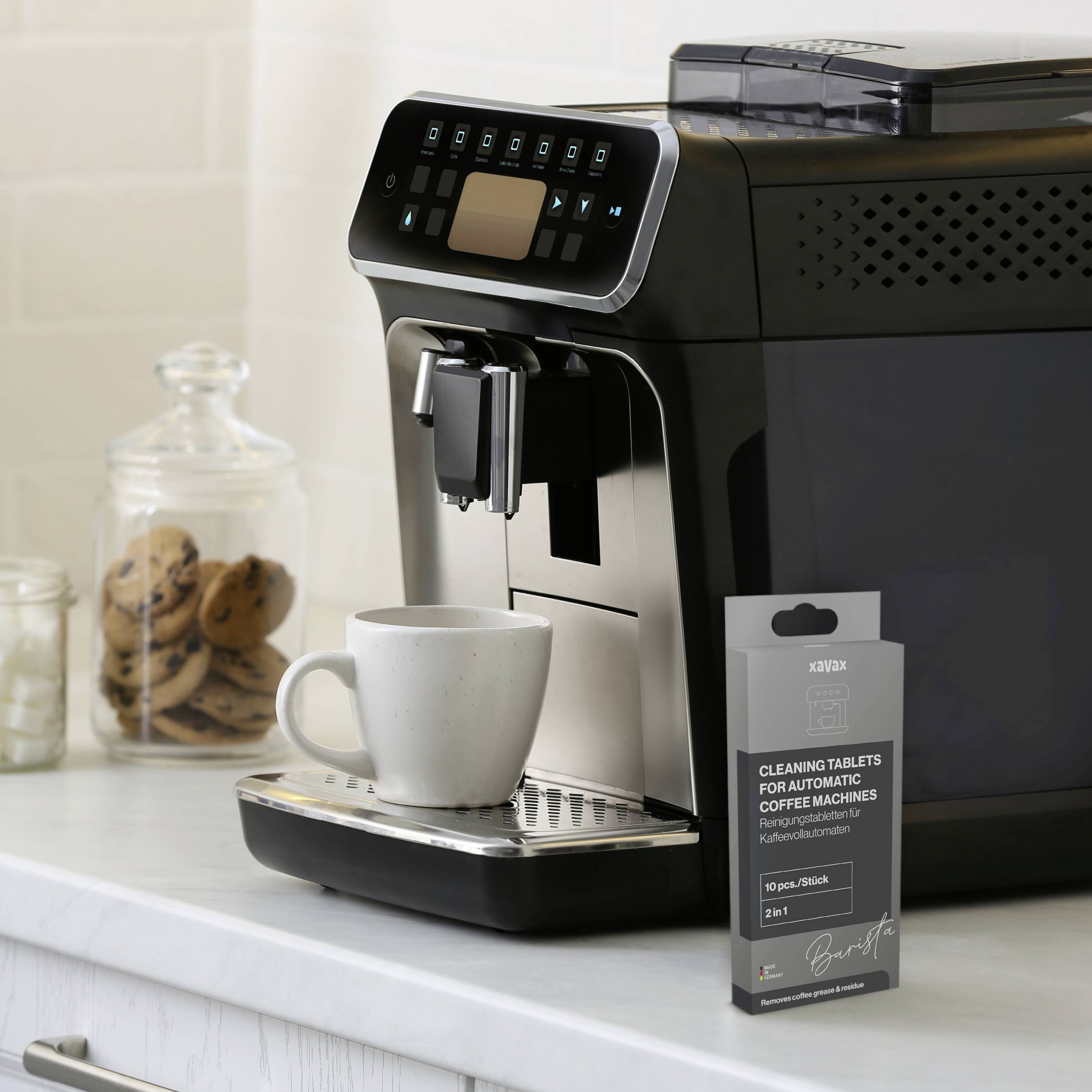 Machine à café noire avec écran posée sur le plan de travail de la cuisine. À côté, un paquet de pastilles de nettoyage et une tasse blanche. En arrière-plan, un bocal à biscuits et du lait.