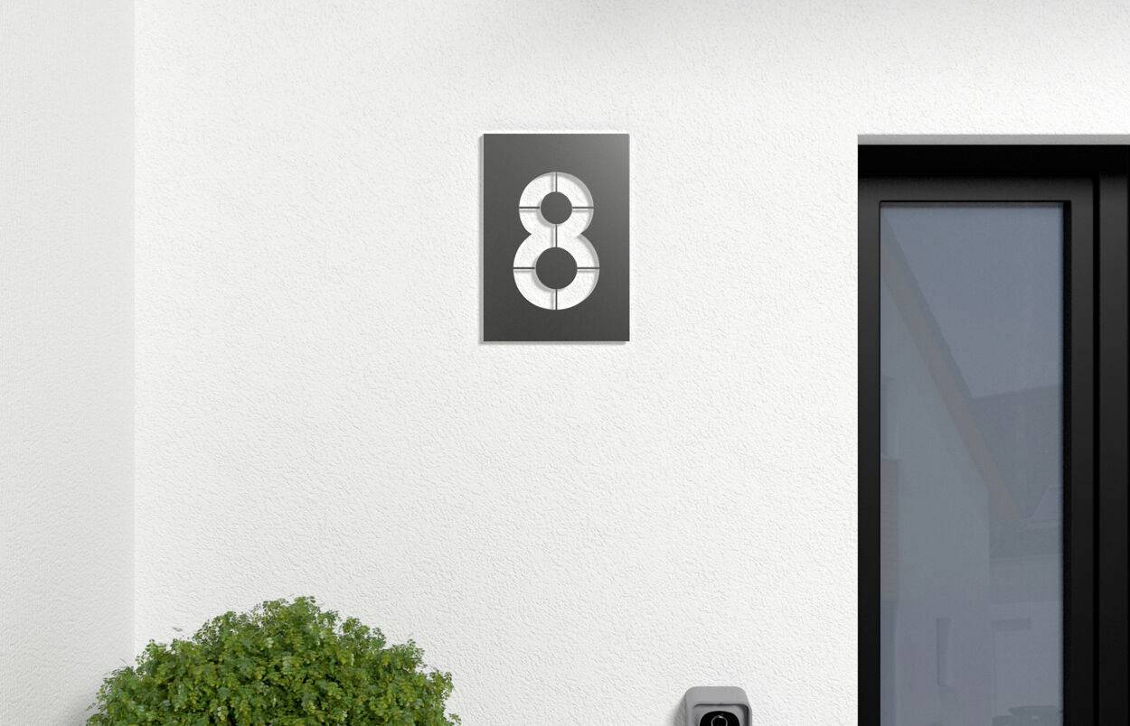 Mur de maison avec le numéro 8 sur une plaque. Une porte noire à côté à droite. En bas à gauche, un buisson vert est visible.